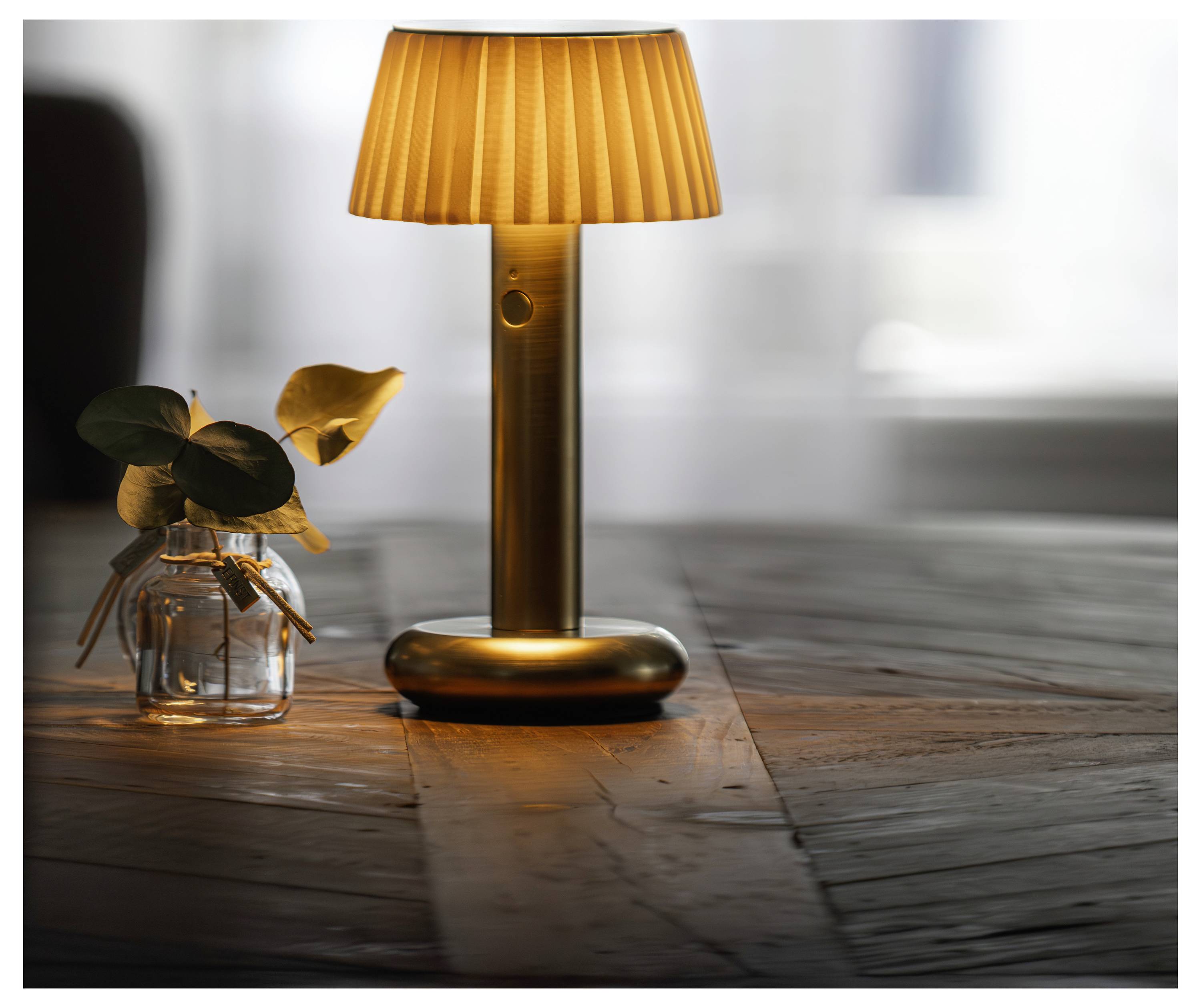 Eine warme bernsteinfarbene Tischlampe leuchtet auf einem Holztisch neben einer kleinen Glasvase mit Blättern.