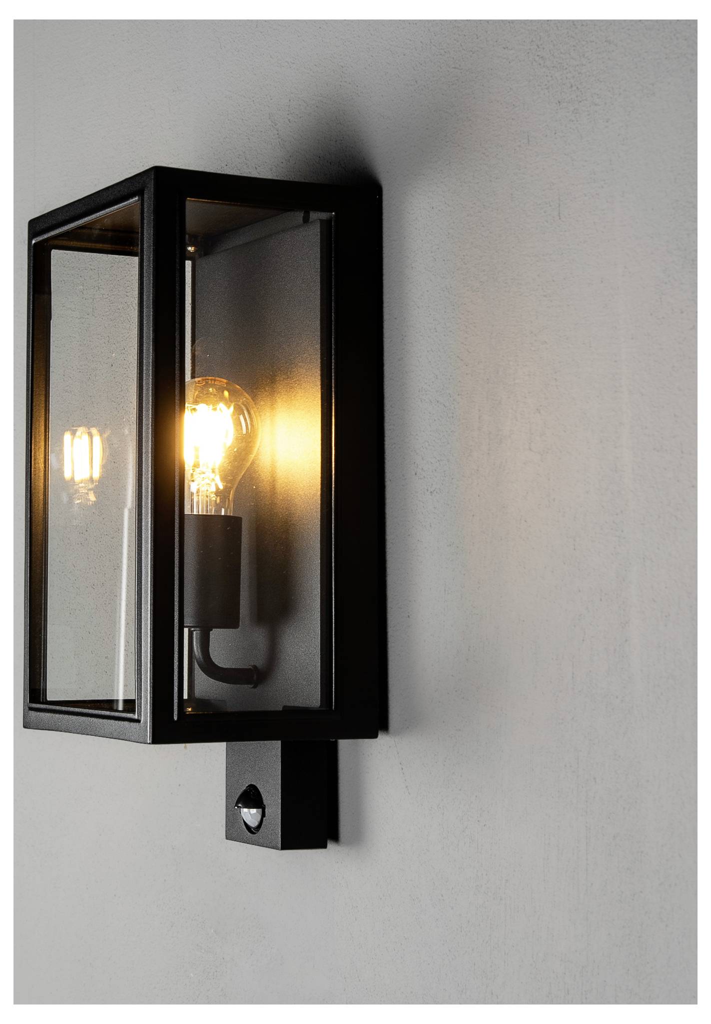 Schwarze Außenwandleuchte mit leuchtender Glühbirne an einer hellgrauen Wand montiert.
