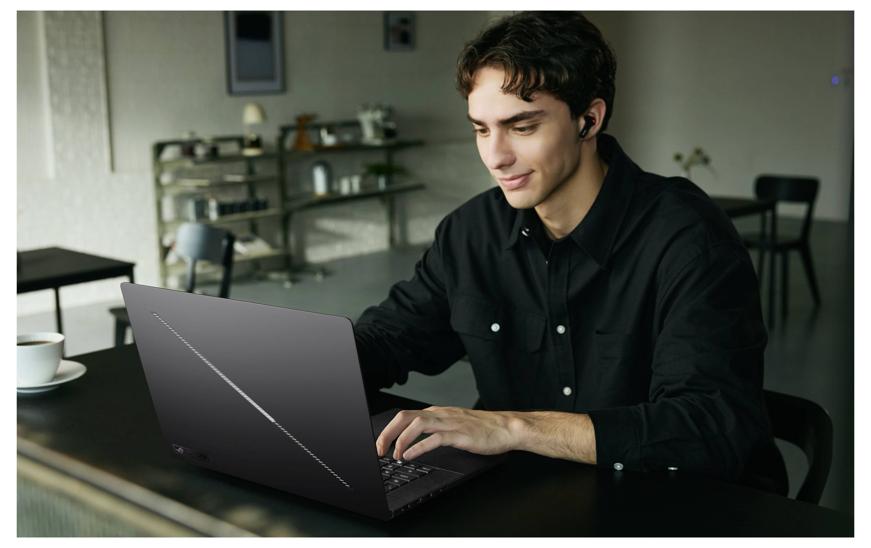 Ein junger Mann in einem dunklen Hemd arbeitet an einem Laptop an einem Tisch in einem modernen Zimmer, trägt Ohrhörer und lächelt leicht.