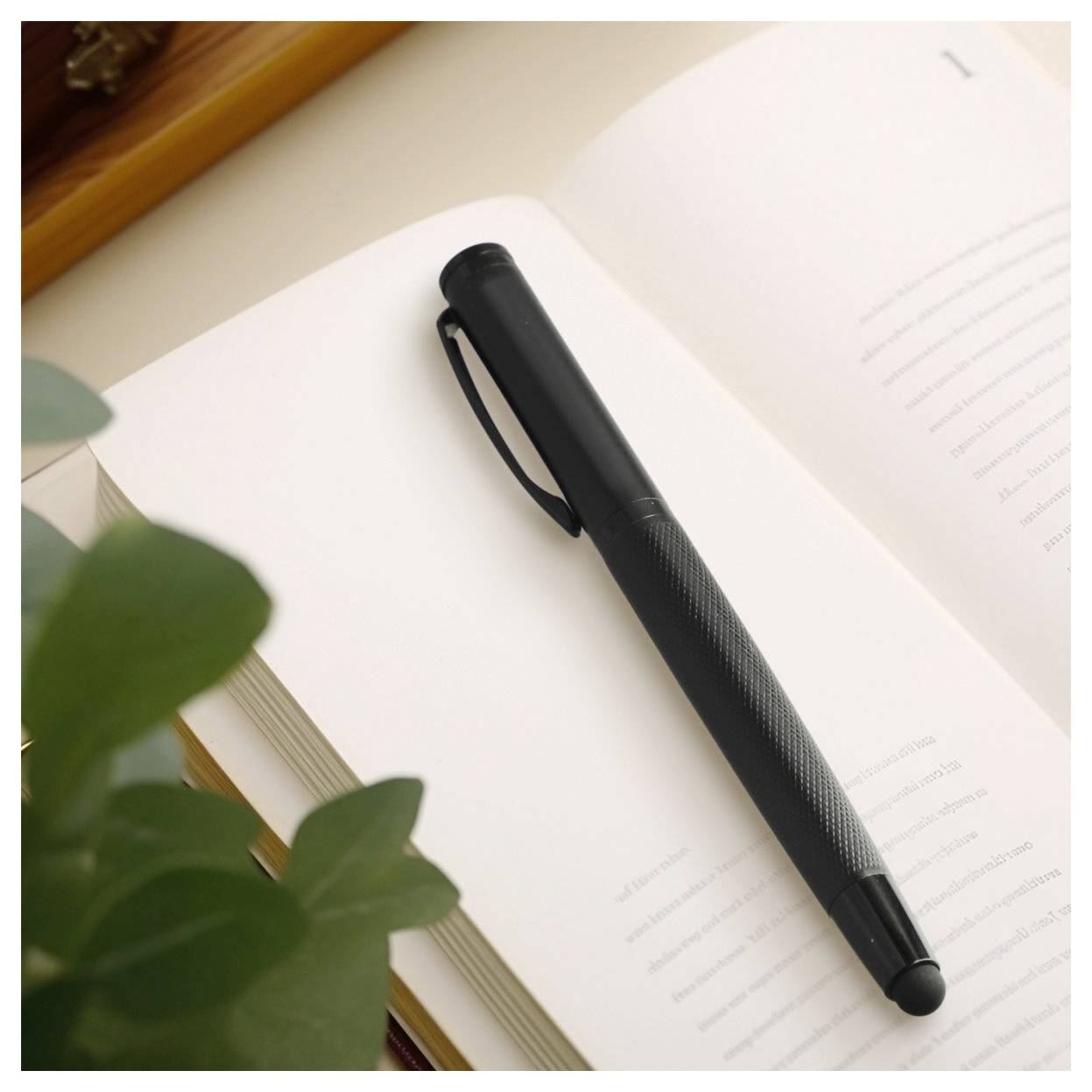 Schwarzer Stift, der diagonal auf einem offenen leeren Notizbuch neben grünen Blättern auf einem Schreibtisch ruht.