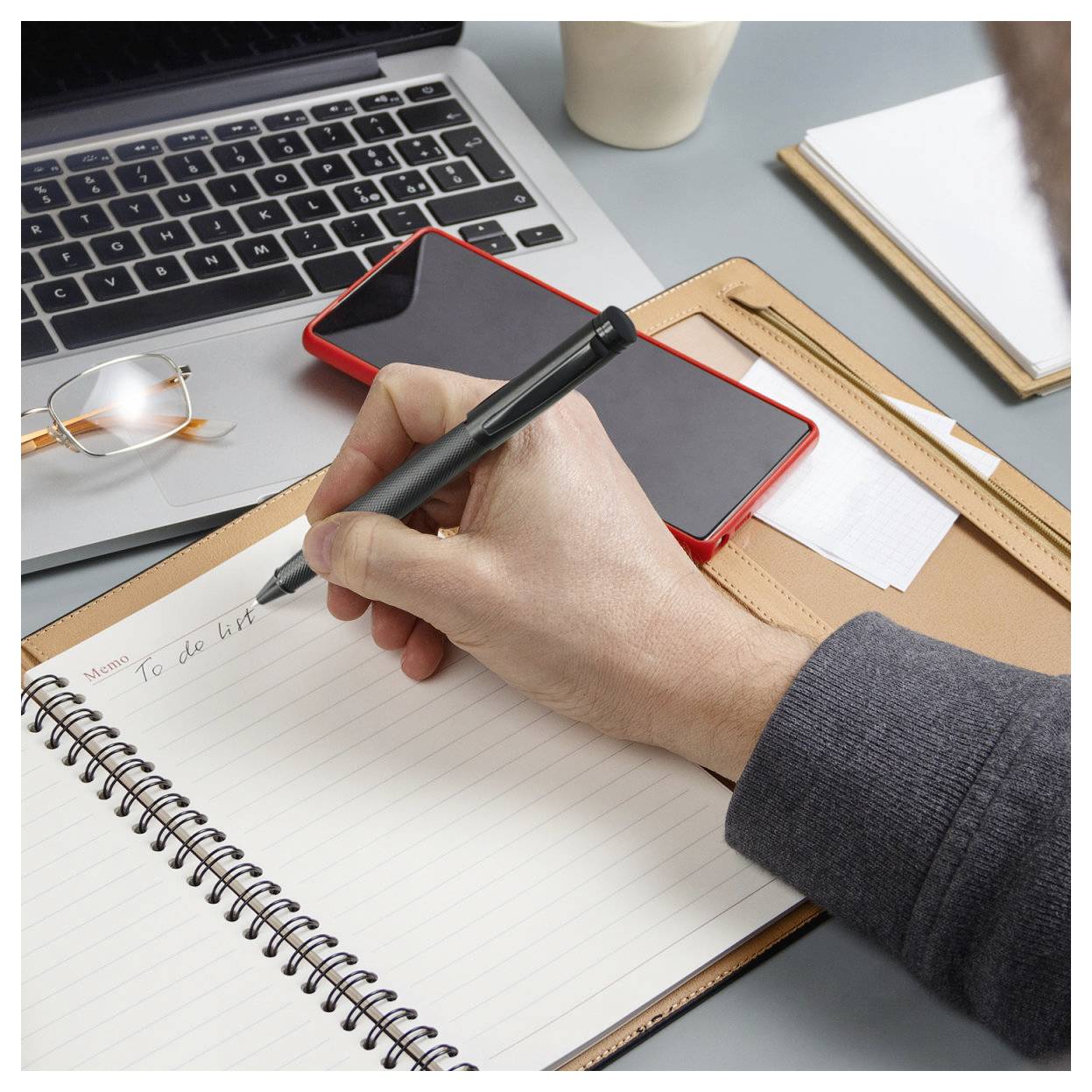 Eine Hand schreibt eine To-Do-Liste in ein Notizbuch neben einem Laptop, Smartphone, Brille und Schreibtischzubehör in einem Büroarbeitsplatz.