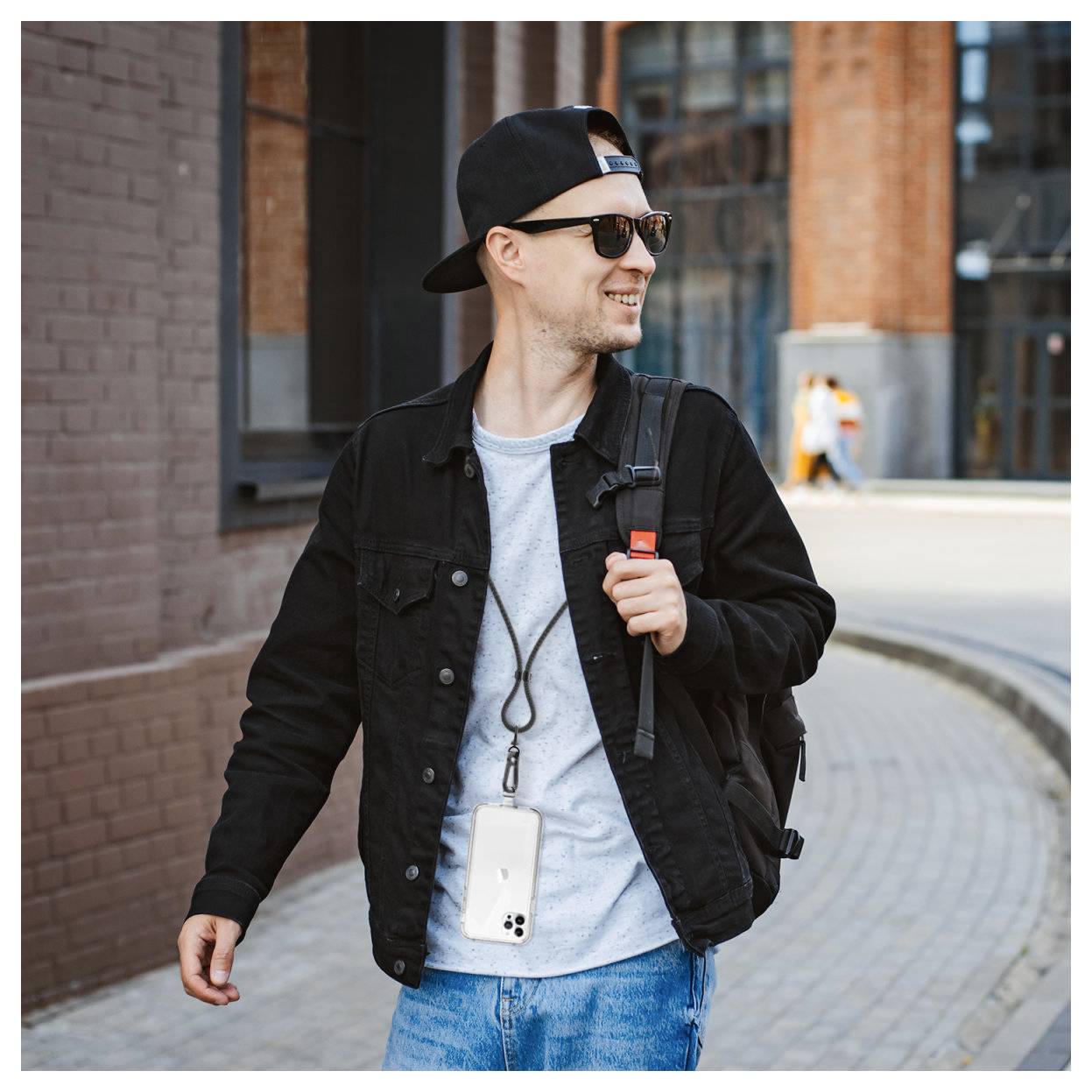 Lächelnder Mann mit Sonnenbrille und rückwärts gedrehter Kappe geht draußen spazieren, trägt eine schwarze Jacke und einen Rucksack mit einem Telefon an einem Umhängeband.