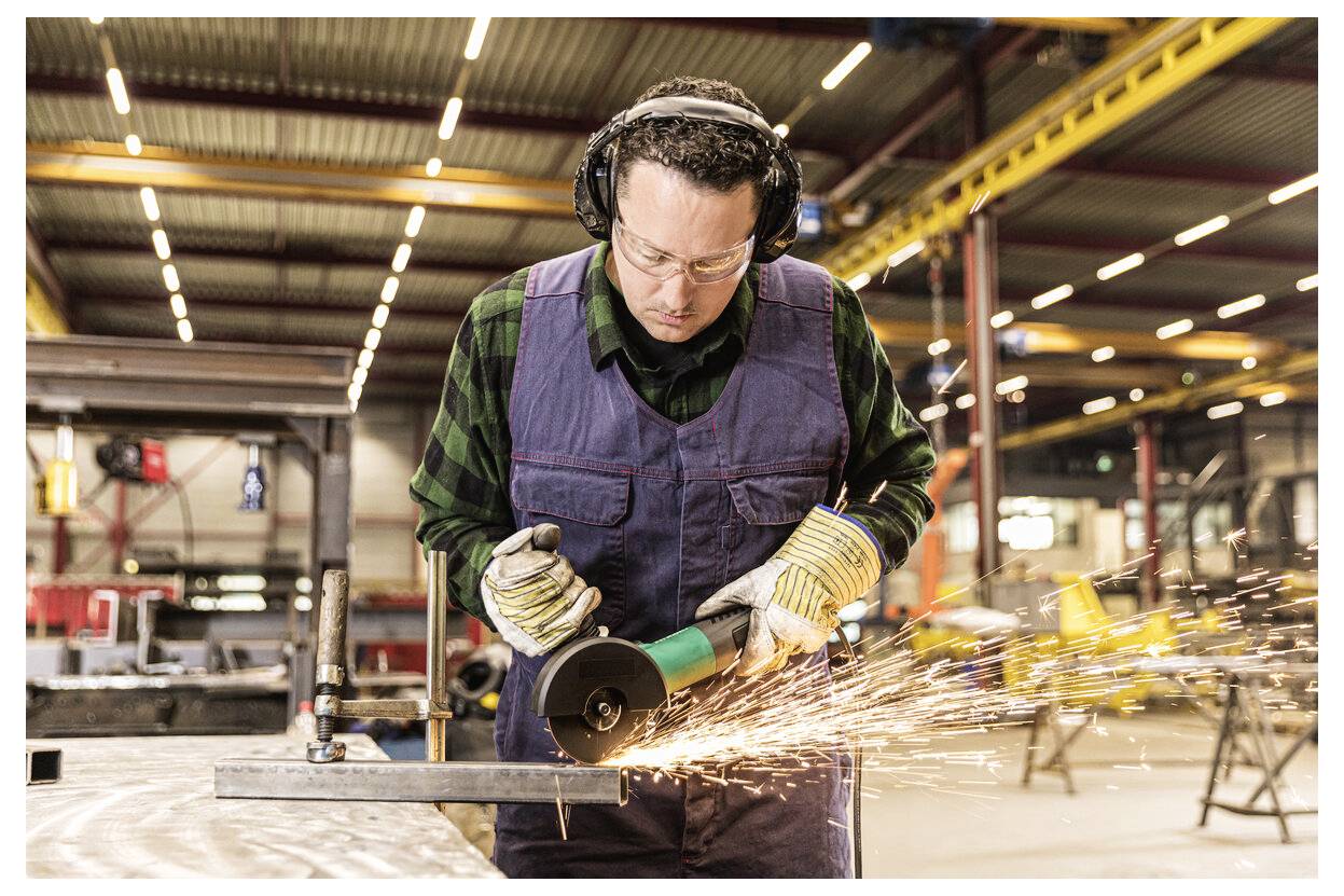 Ein Arbeiter in Schutzbrille und Gehörschutz benutzt einen Winkelschleifer auf Metall in einer Fabrik, wobei Funken über die Werkstatt fliegen.