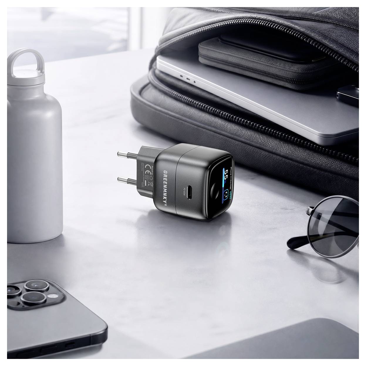 Schwarzes USB-C-Wandladegerät mit digitalem Display neben einer Laptoptasche, einem Telefon, einer Sonnenbrille und einer Wasserflasche auf einem Schreibtisch