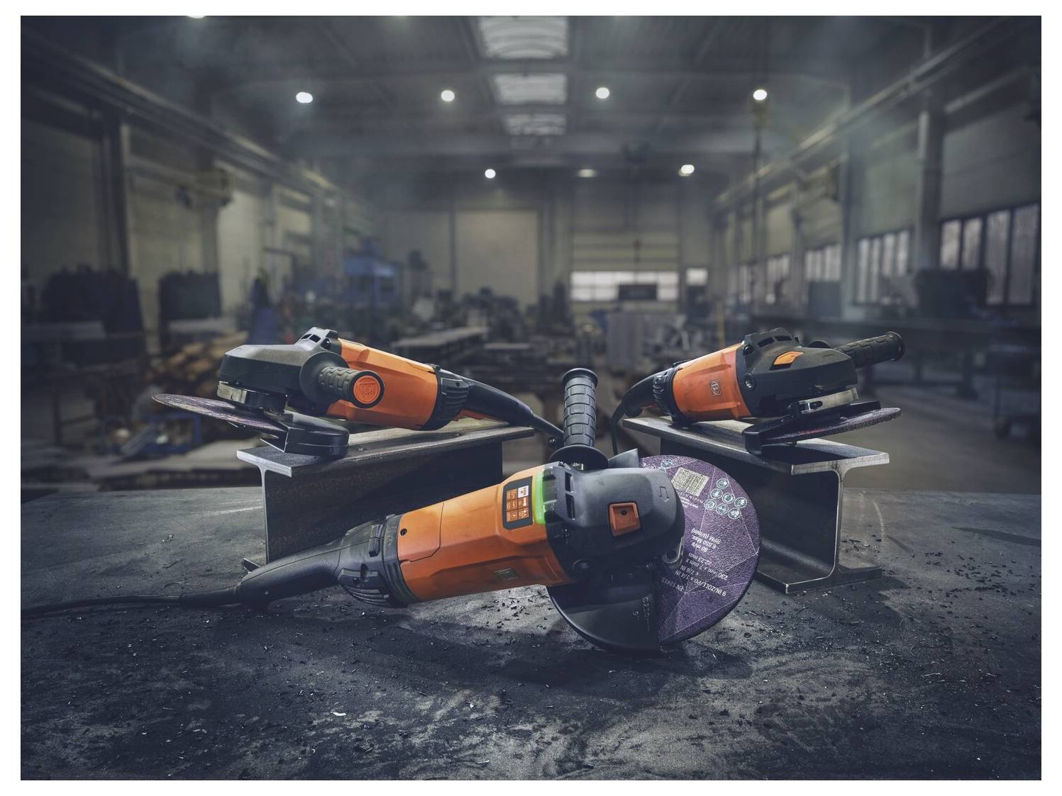 Drei orange-schwarze Elektrowerkzeuge liegen auf einem Betonboden in einer Werkstatt, mit einem großen Winkelschleifer vorne und einem industriellen Garagenhintergrund.