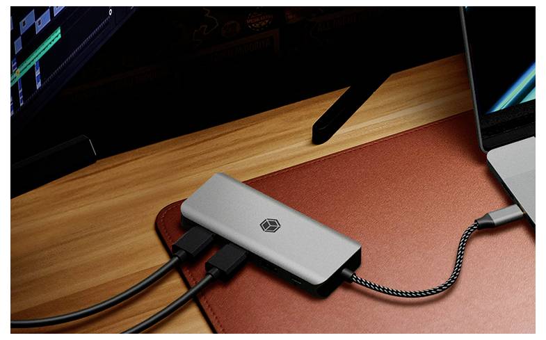 USB-C-Hub auf einem Schreibtisch, der einen Laptop und Zubehör verbindet, mit mehreren Kabeln, die in den kompakten silbernen Adapter auf einer roten Matte eingesteckt sind.