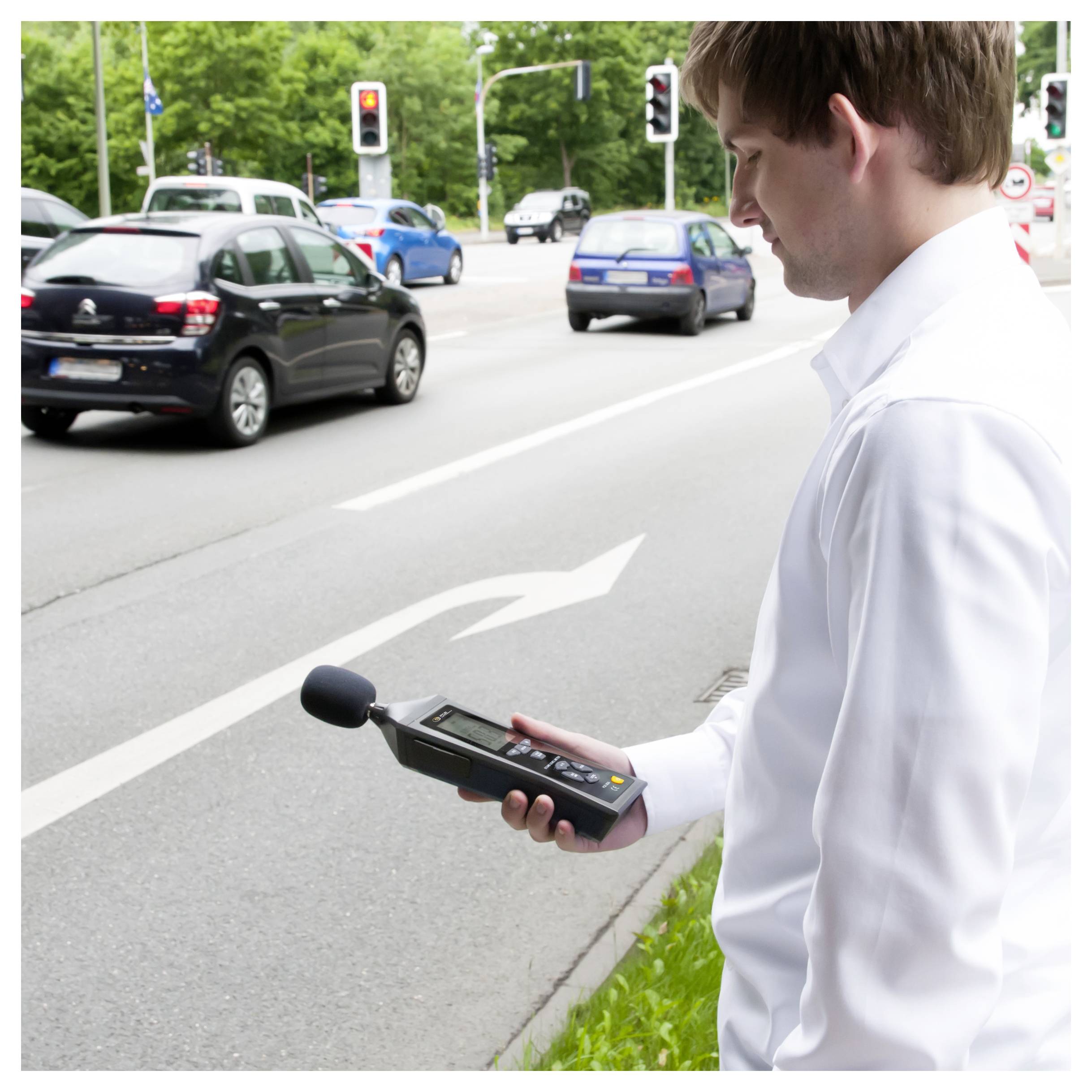 Mann im weißen Hemd, der ein tragbares Schallpegelmessgerät neben einer belebten Straße an einer Ampel verwendet und Verkehrslärm misst.