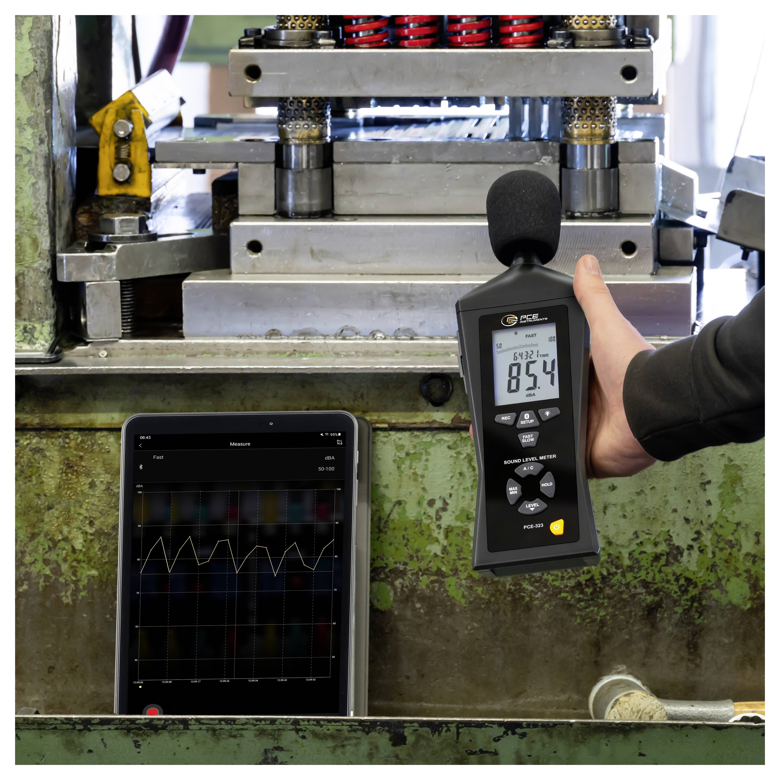 Eine Person misst Industriemaschinenlärm mit einem tragbaren Dezibelmessgerät, während ein Tablet ein Schallpegel-Diagramm anzeigt.