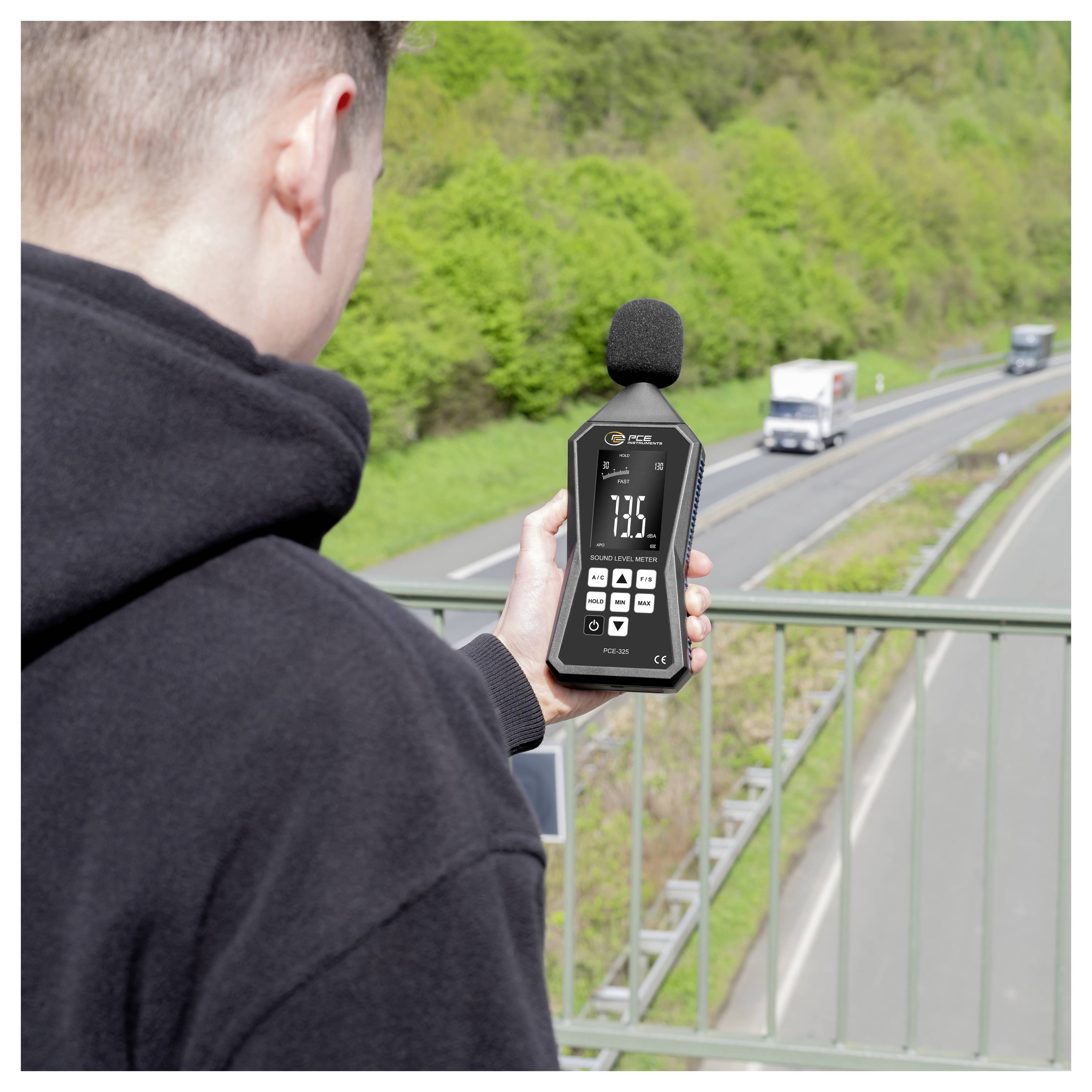 Person, die ein Schallpegelmessgerät mit Schaumstoff-Mikrofon hält, misst Straßenverkehrslärm von einer Überführung aus.