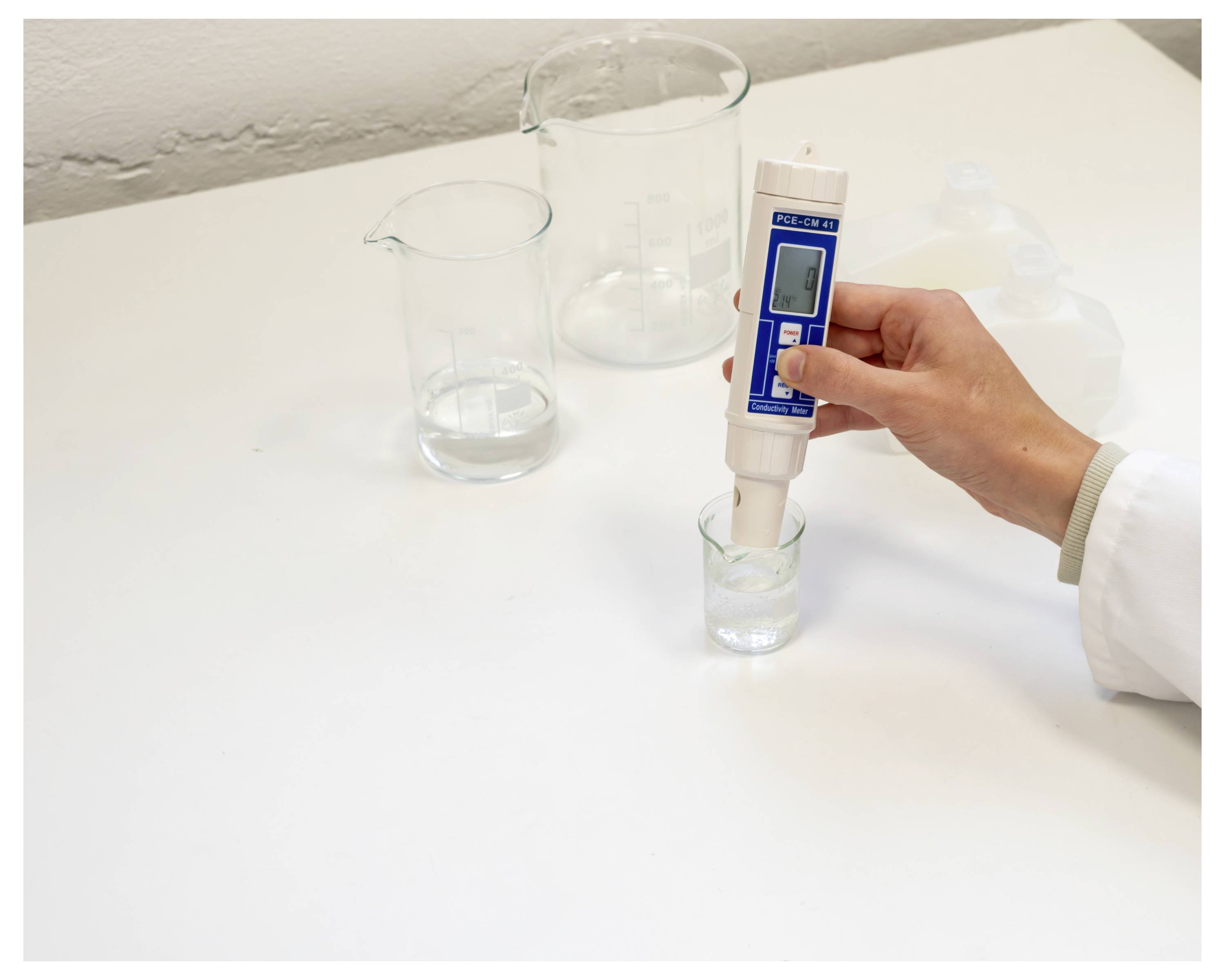 Eine Hand hält ein digitales Wassergütemessgerät in einem kleinen Becher und misst Flüssigkeit in einer Laboraufstellung mit größeren Bechern in der Nähe.