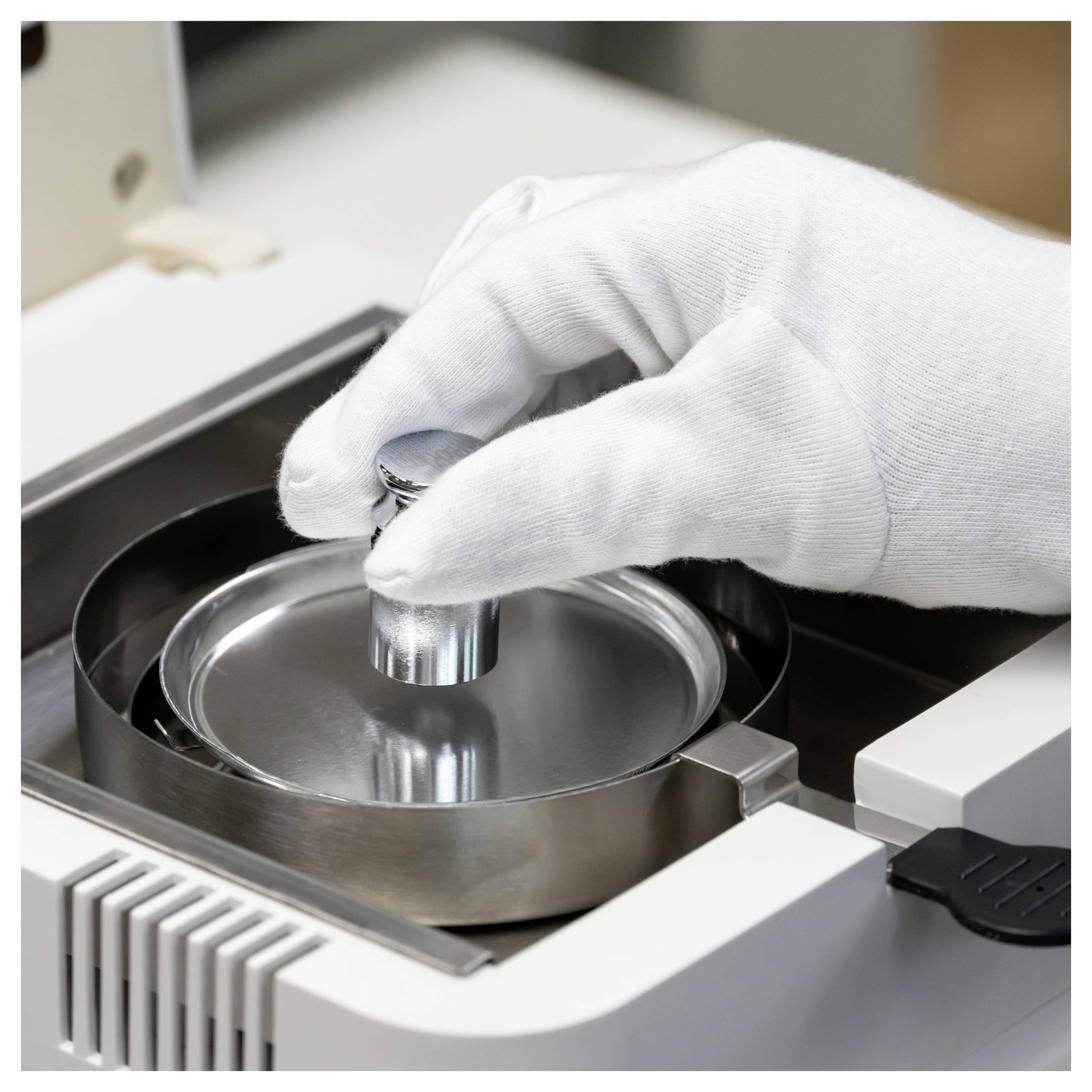 Eine behandschuhte Hand legt eine Probe auf die kreisförmige Metalloberfläche eines Laborinstruments zur Messung oder Prüfung.