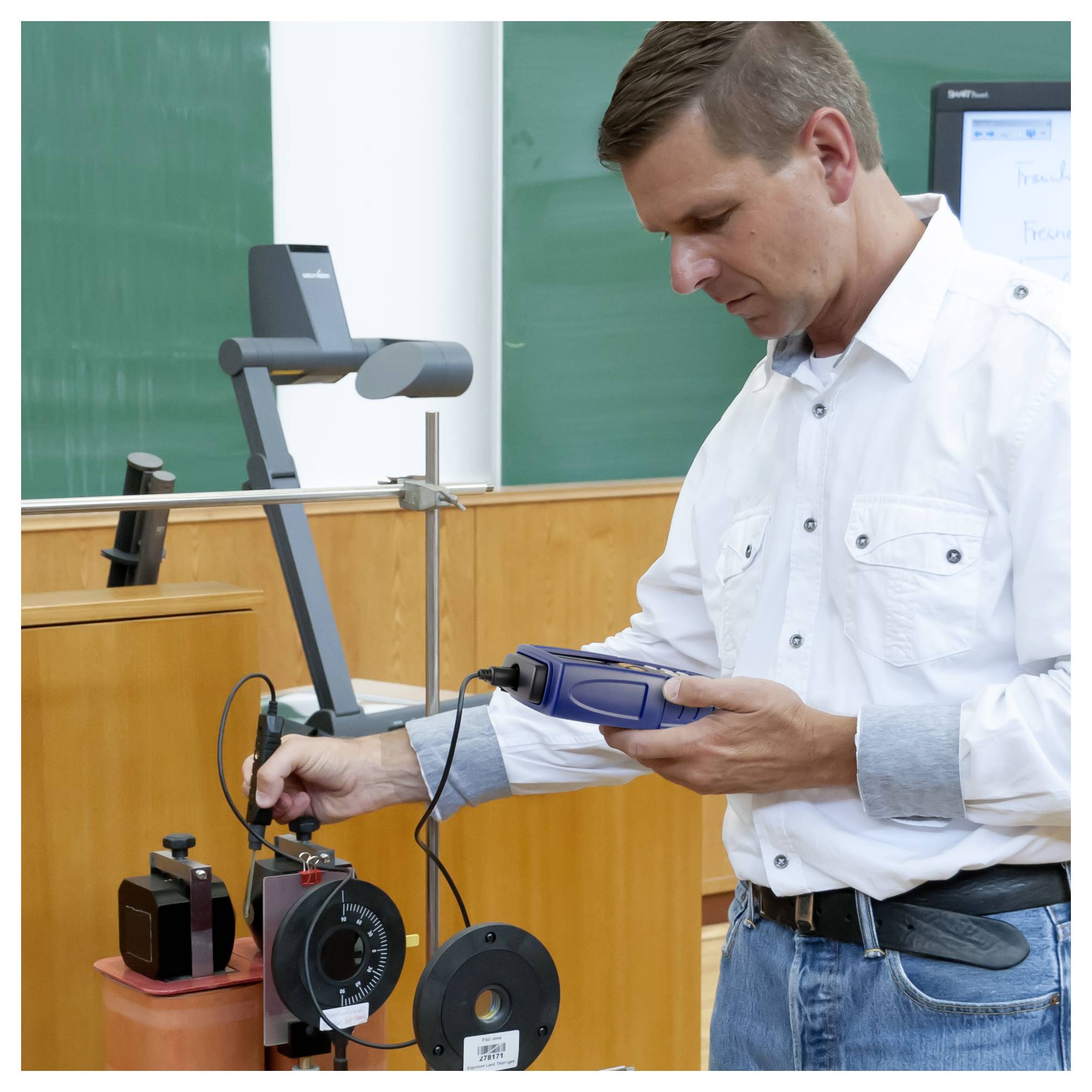 Mann in einem Klassenzimmer, der ein tragbares Gerät bedient, das mit Laborausrüstung und einer Spule verbunden ist, was auf eine technische Demonstration oder ein Experiment hindeutet.
