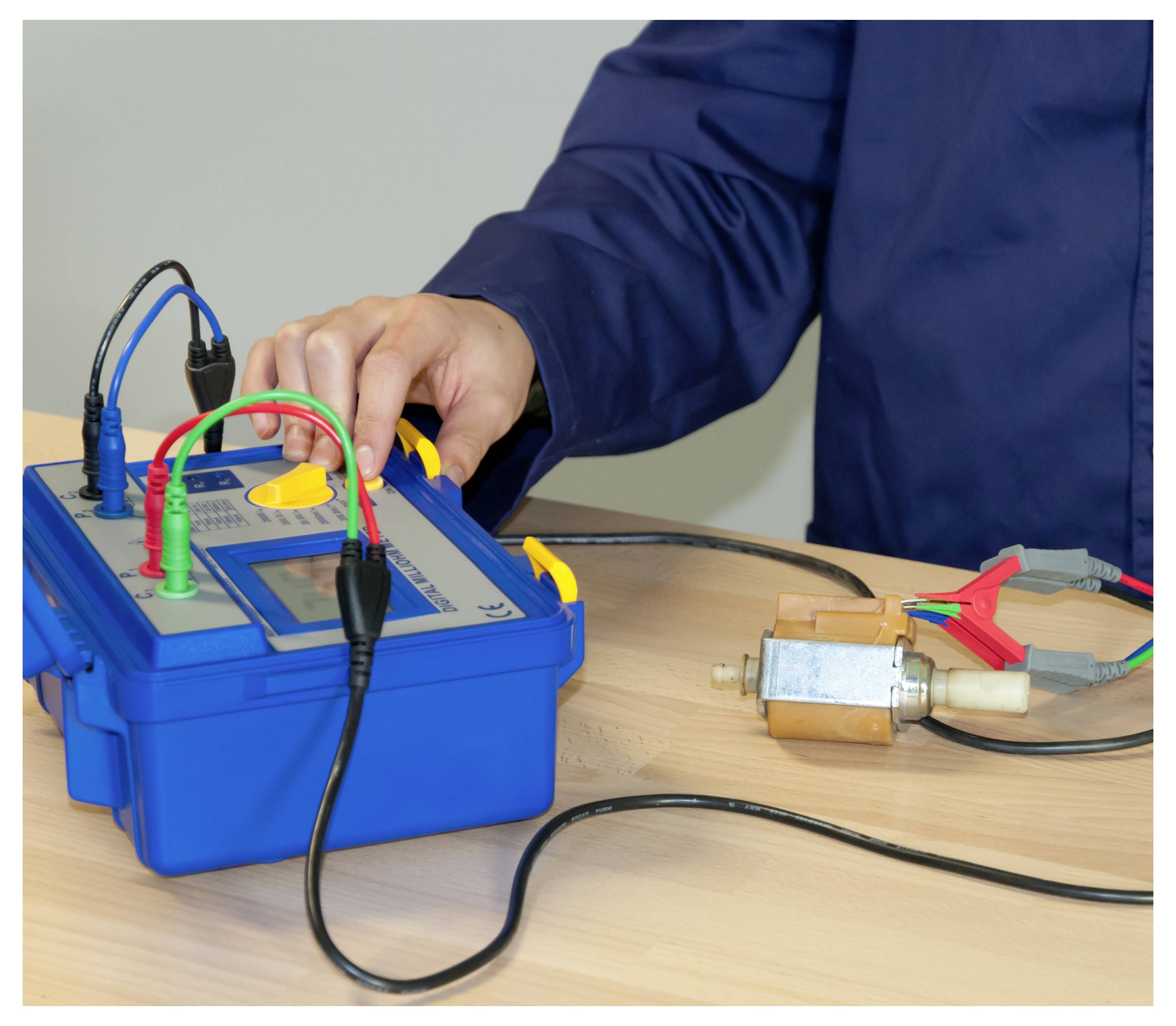 Person, die ein elektrisches Gerät mit einer blauen Diagnose-Box und Multimeter-Leitungen testet, die mit einem Motor auf einem Tisch verbunden sind.