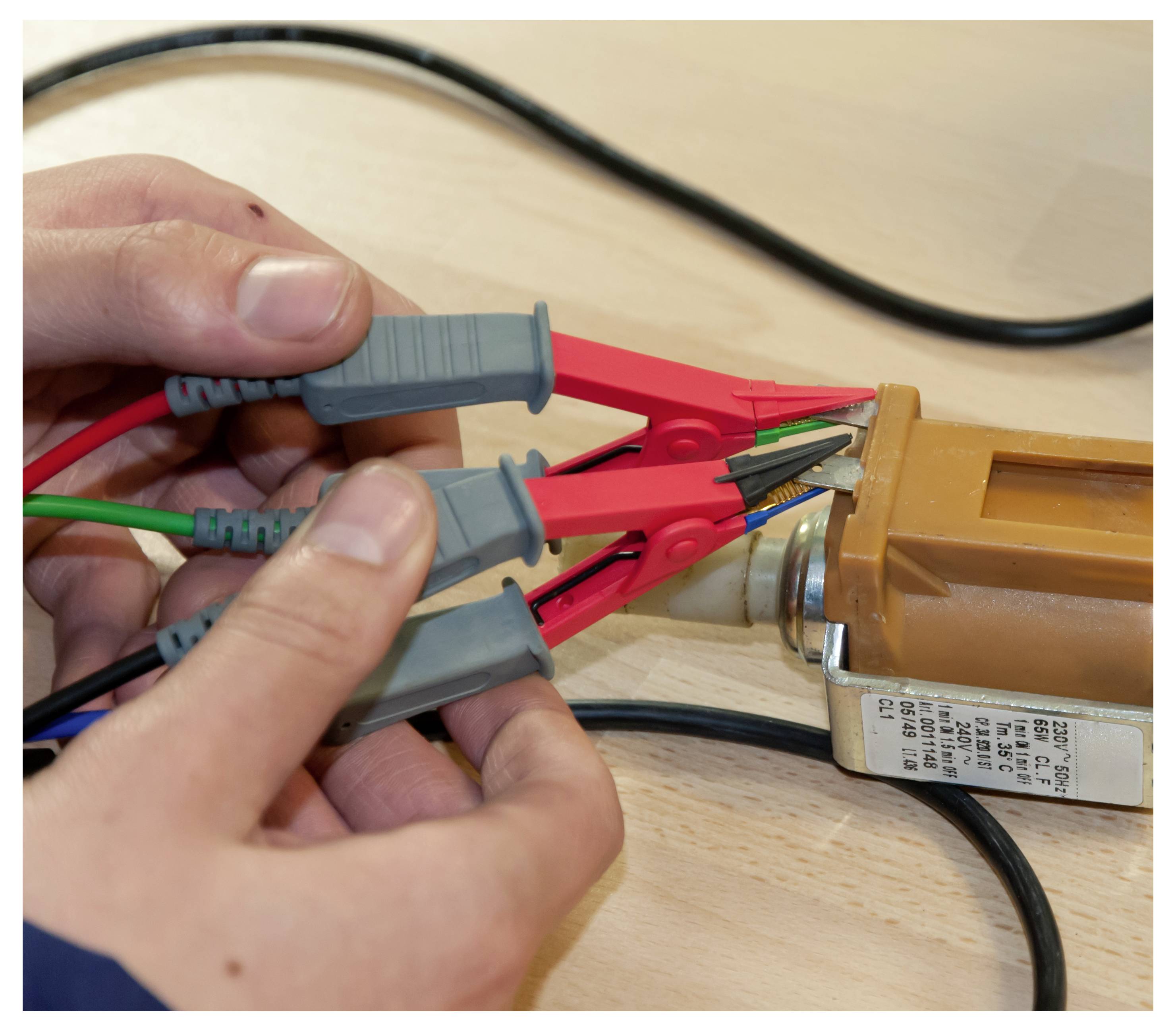 Hände halten drei Messleitungen mit roten Krokodilklemmen, die an einer elektrischen Komponente auf einer Holzoberfläche befestigt sind.