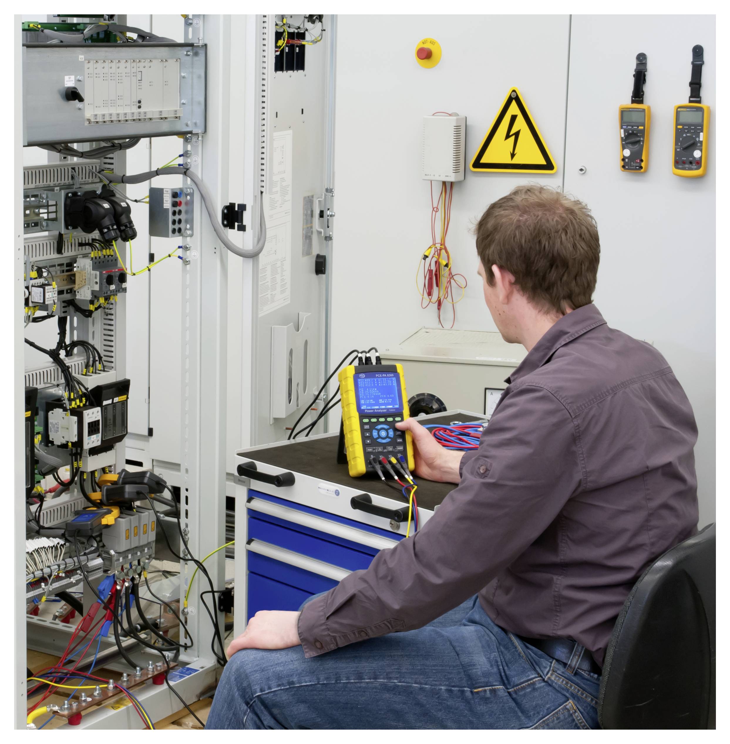 Techniker testet Elektrosteuerpanelverkabelung mit einem tragbaren Messinstrument in einer Werkstatt neben einem Hochspannungswarnschild.