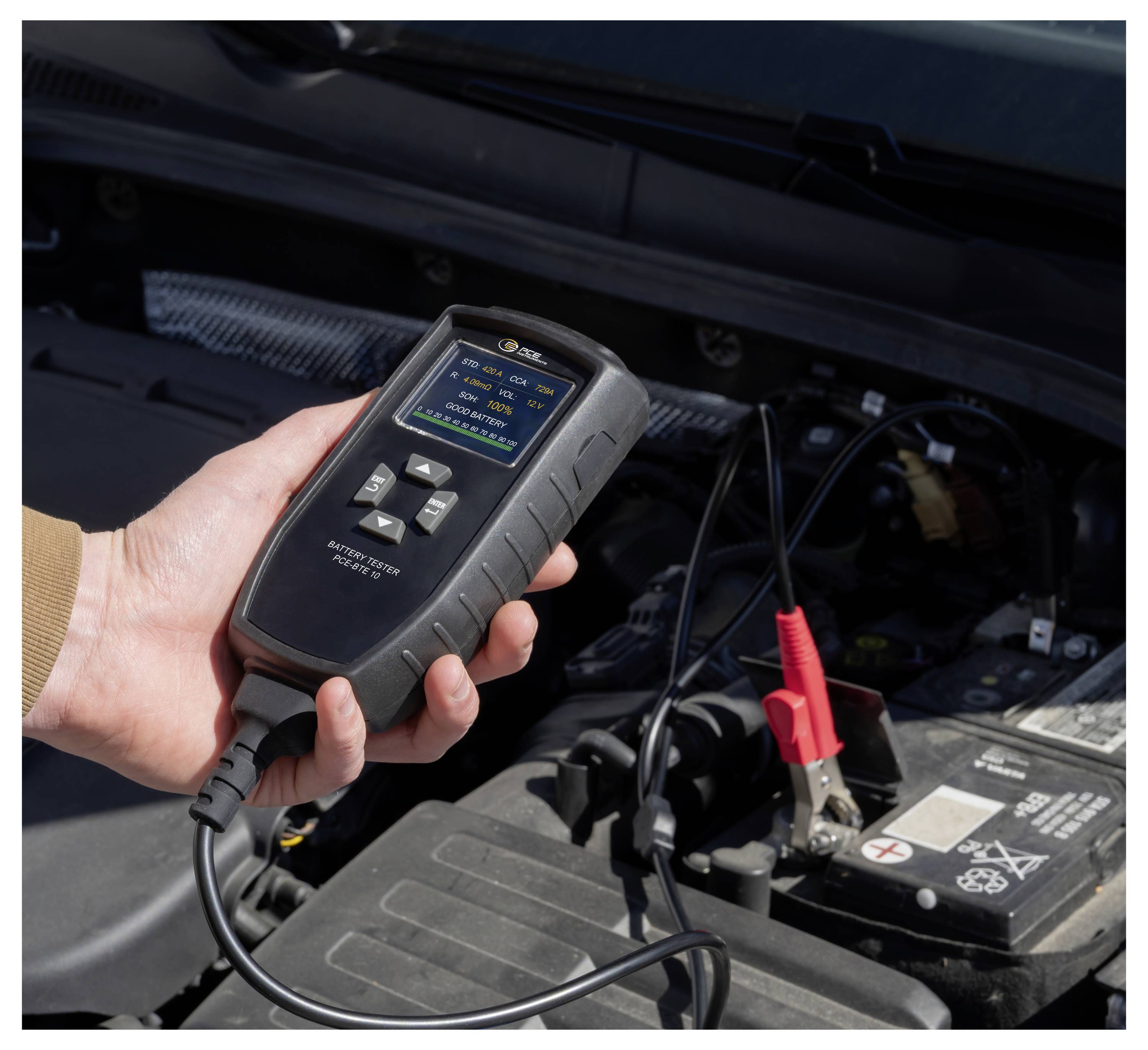 Hand hält einen Batterietester in der Hand, der mit einer Autobatterie unter der Motorhaube verbunden ist, um den Status der Fahrzeugbatterie zu überprüfen.
