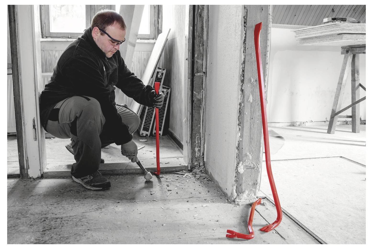 Ein Mann benutzt eine Brechstange, um Schutt in einer Türöffnung in einem unfertigen Raum zu entfernen, mit roten Sicherheitsmarkierungen auf dem Boden und der Wand.