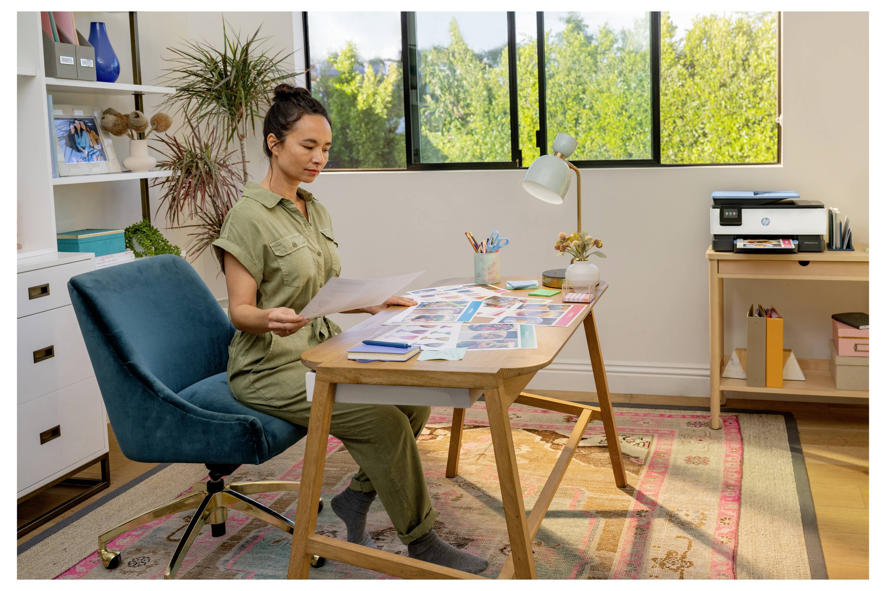 Eine Frau sitzt an einem Schreibtisch und überprüft gedruckte Dokumente in einem hellen Arbeitszimmer zu Hause, umgeben von Papieren, einer Lampe, einem Stuhl und Regalen.