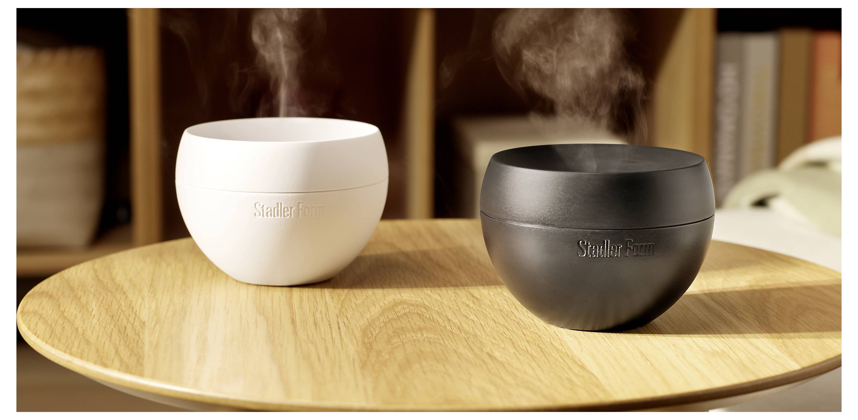 Zwei kleine schalenförmige Luftbefeuchter, einer weiß und einer schwarz, geben Dampf auf einem Holztablett ab.