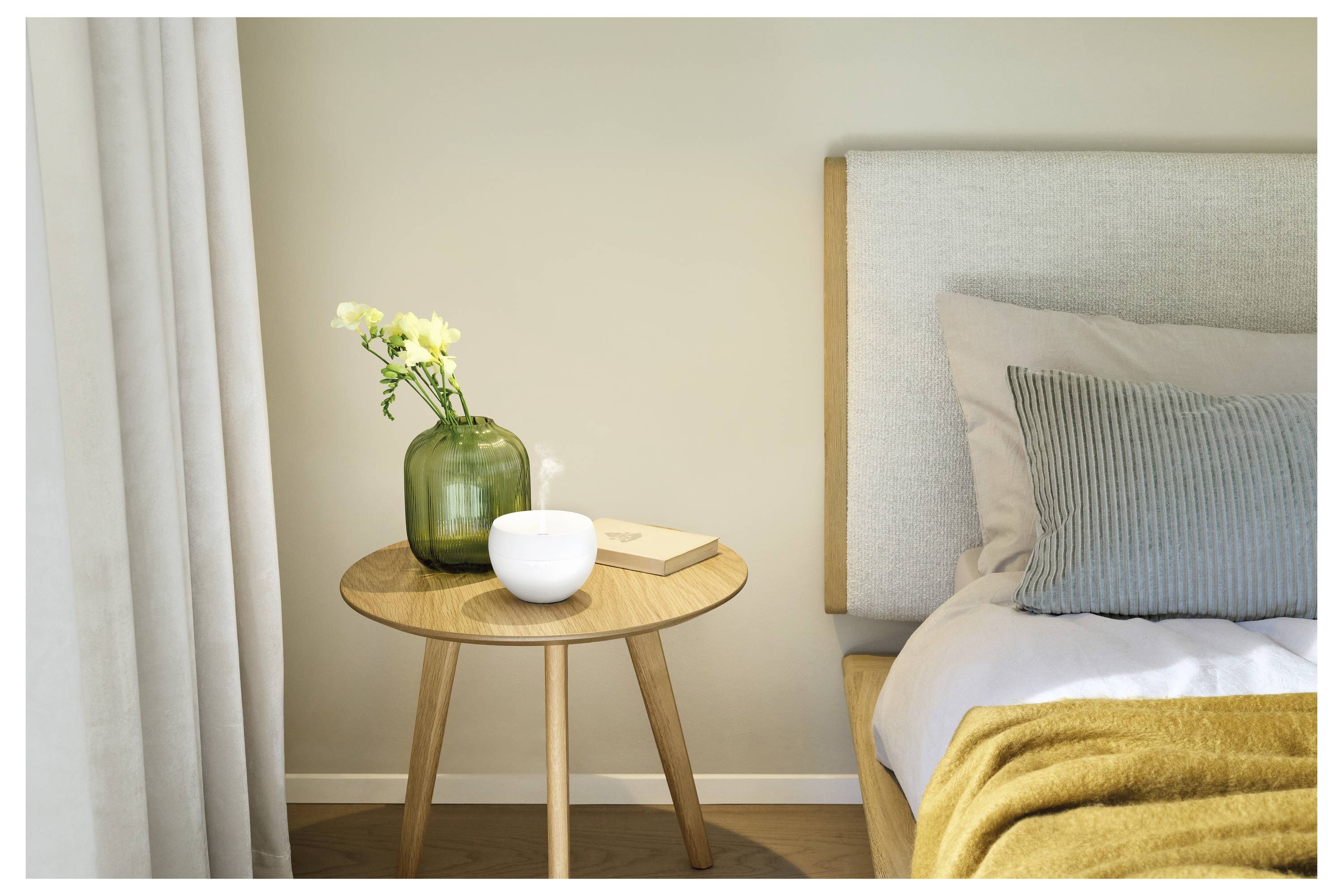 Moderne Schlafzimmerecke mit einem Nachttisch, auf dem eine grüne Vase mit Blumen, eine weiße Schüssel und Bücher neben einem ordentlich gemachten Bett mit Kissen und einer senfgelben Wurfdecke stehen.