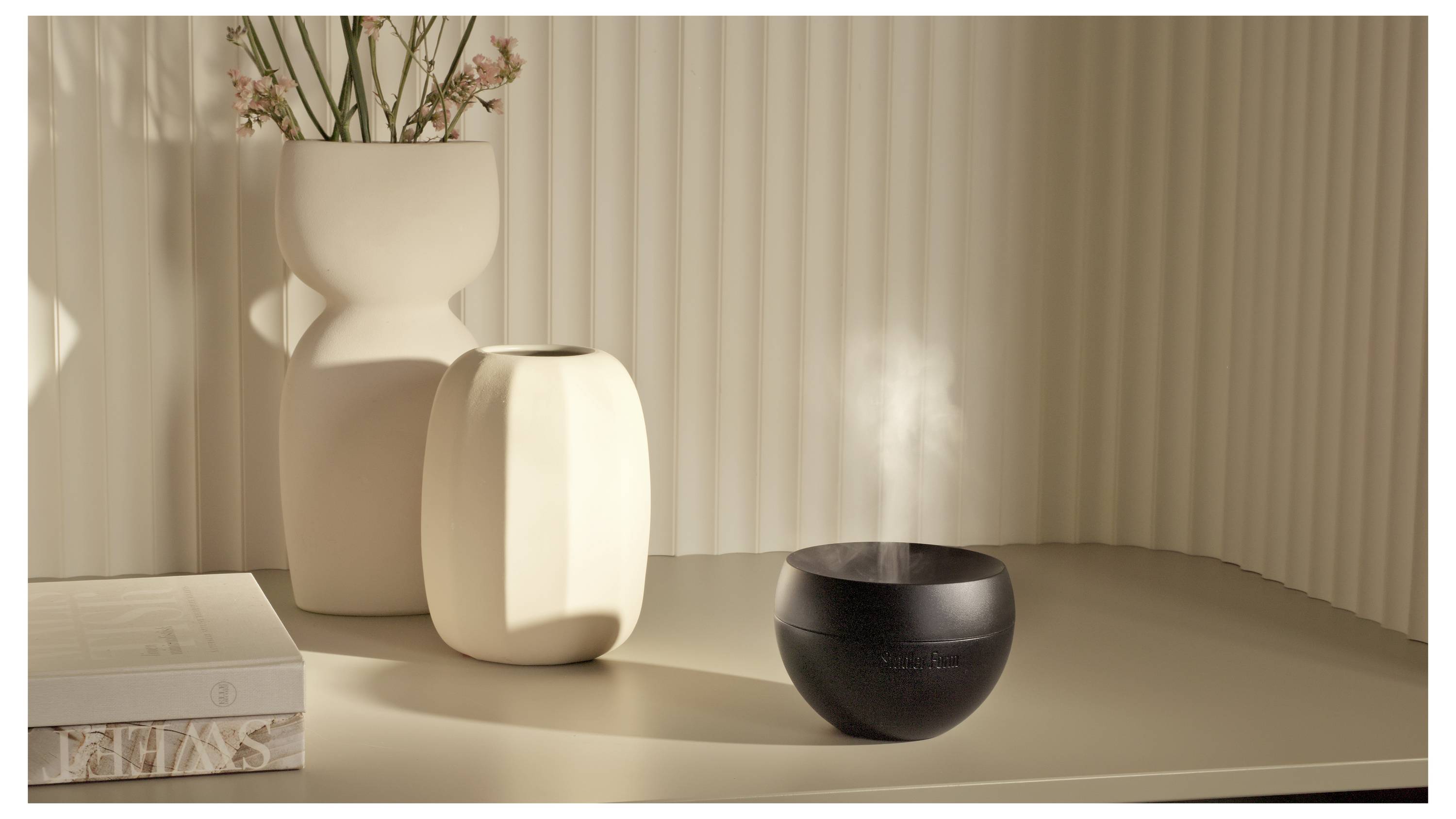 Eine schwarze, schalenförmige Duftlampe verströmt Dampf auf einer beigefarbenen Tischoberfläche neben dekorativen Vasen und Büchern im warmen Sonnenlicht.