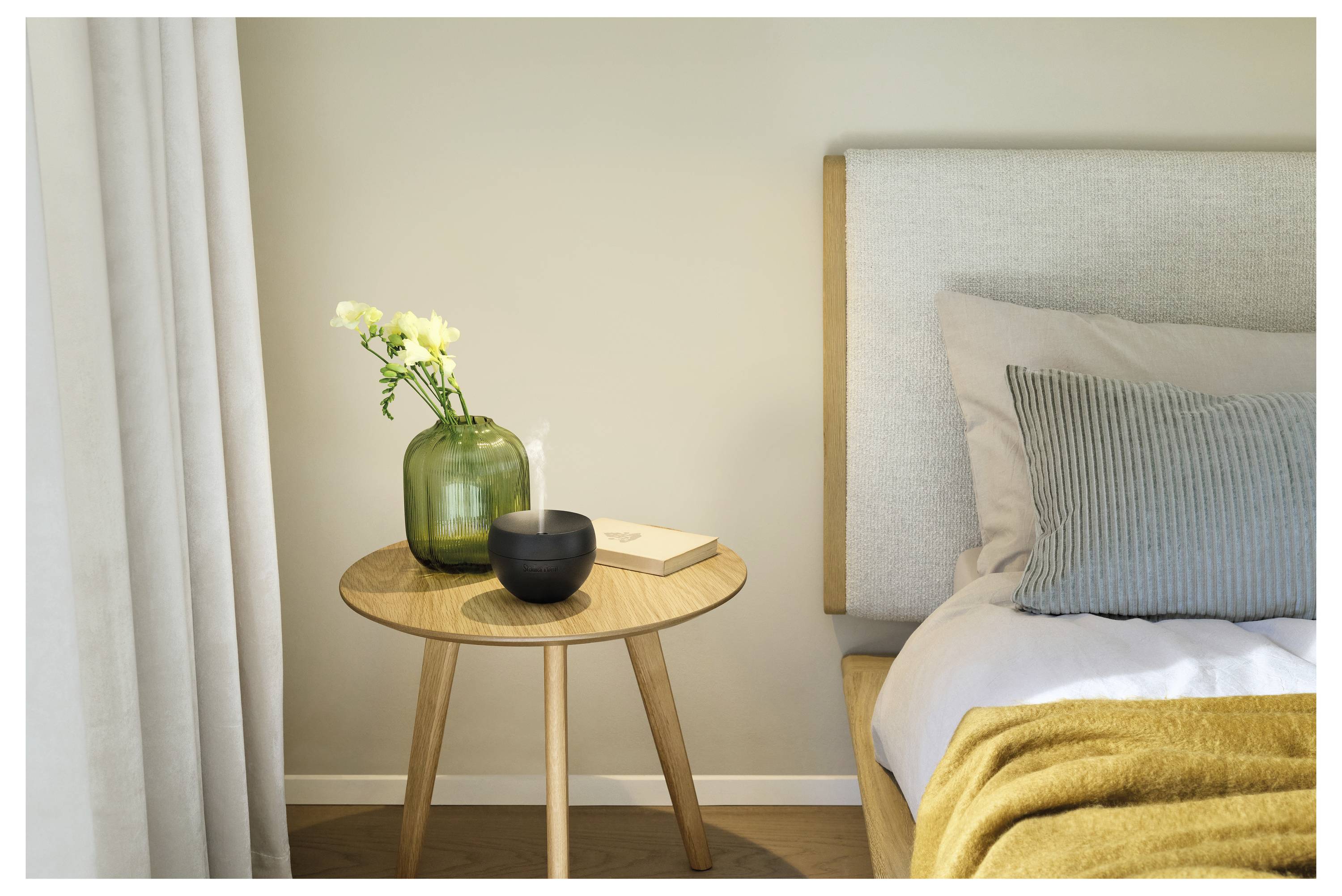 Ein ordentlich gemachtes Bett neben einem kleinen Holznachttisch mit einer grünen Vase, Blumen, einer Schale und einem Buch in einem ruhigen Schlafzimmer.
