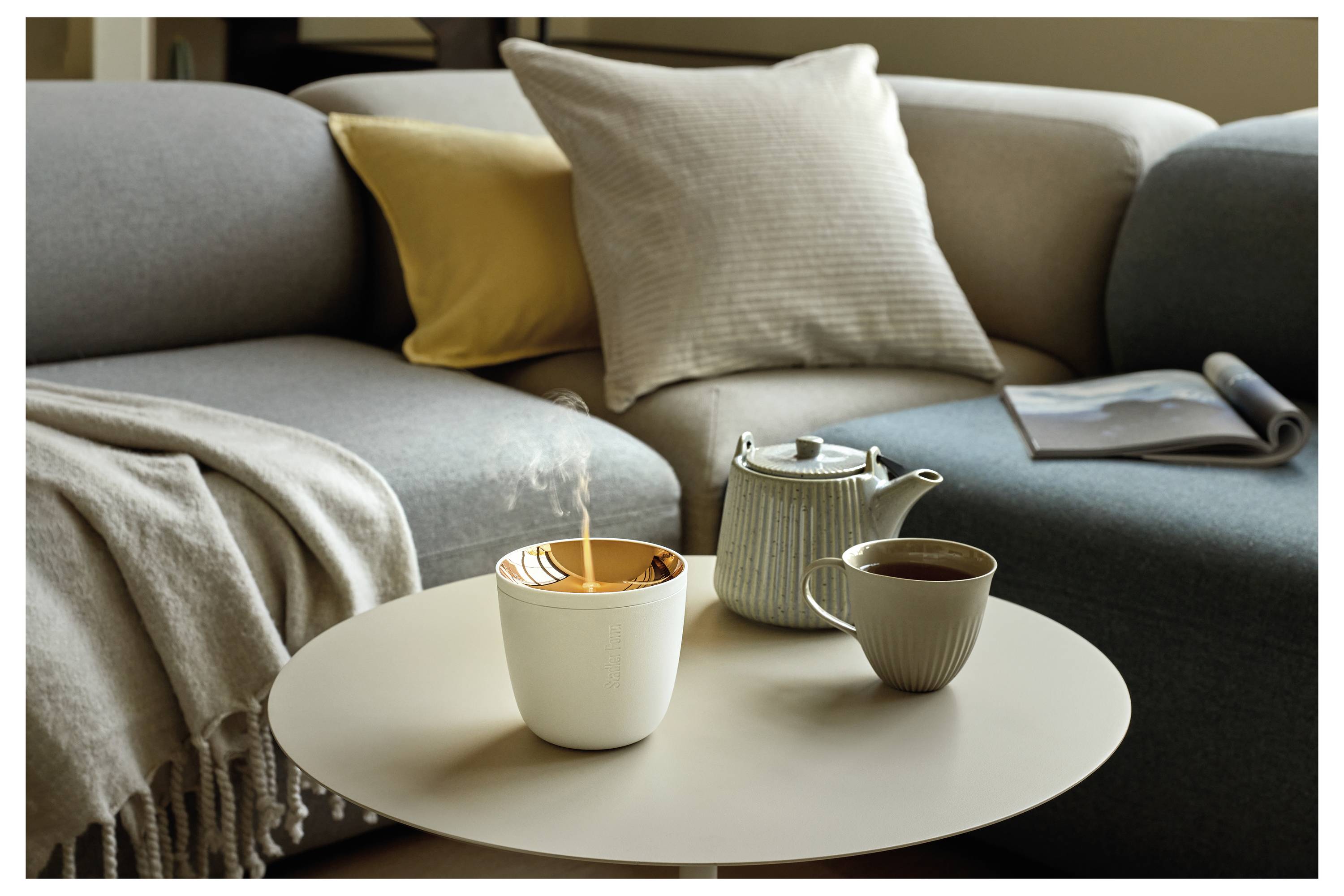 Gemütliche Wohnzimmerszene mit einem grauen Sofa, Kissen, Wurfdecke und rundem Couchtisch, auf dem eine brennende Kerze, eine Teekanne und eine Tasse stehen.