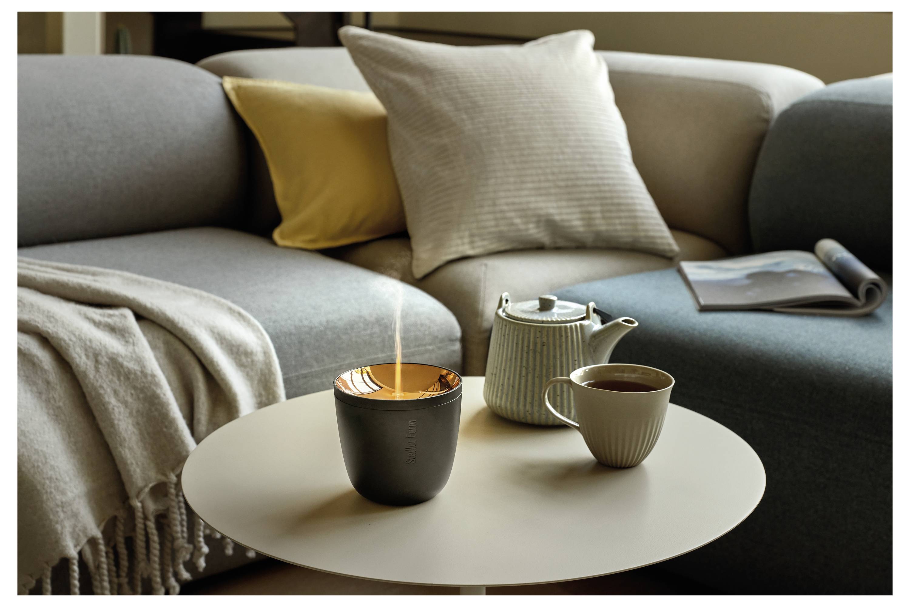 Gemütliche Wohnzimmerszene mit einem grauen Sofa, einer Wurfdecke und Kissen sowie einem runden Couchtisch, auf dem eine brennende Kerze, eine Teekanne und eine Tasse stehen.