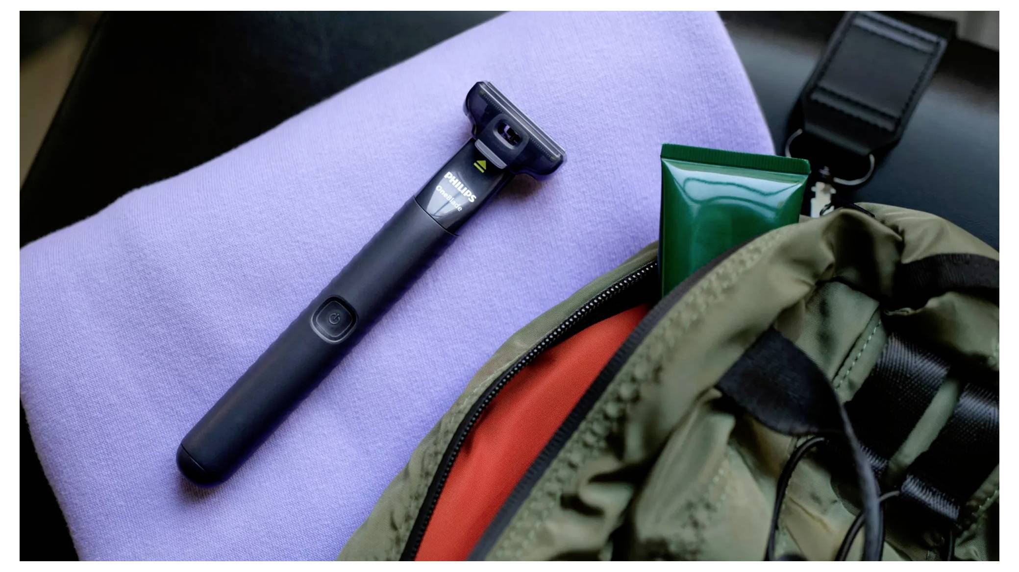 Schwarzer Elektrorasierer auf einem lavendelfarbenen Handtuch neben einem Rucksack, schwarzem Riemen und grüner Tube, was auf ein Reise-Pflege-Set hindeutet.