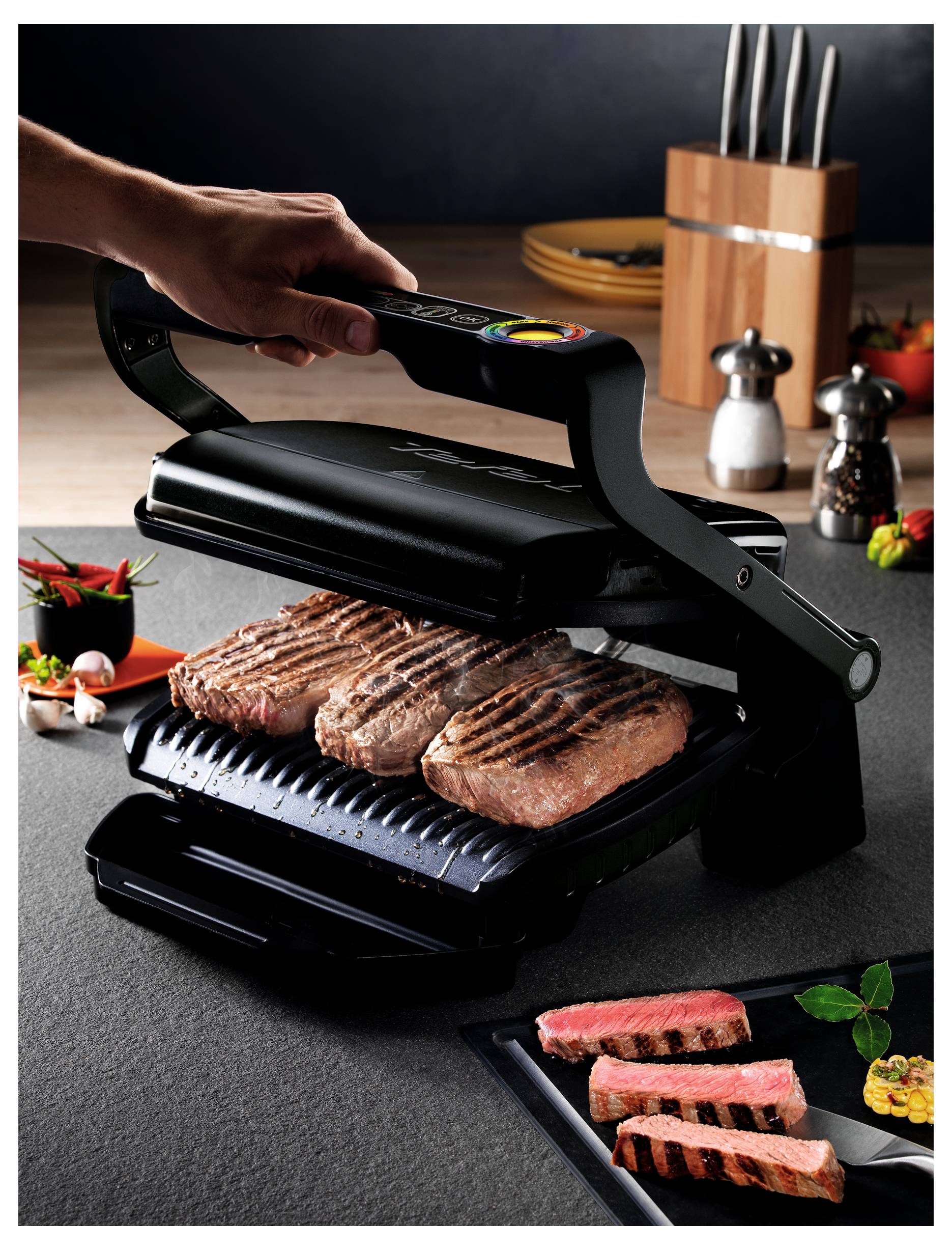 Hand drückt einen Kontaktgrill über drei Steaks zu, mit geschnittenem gegrilltem Rindfleisch, das auf einem Teller daneben in einer Küche serviert wird.