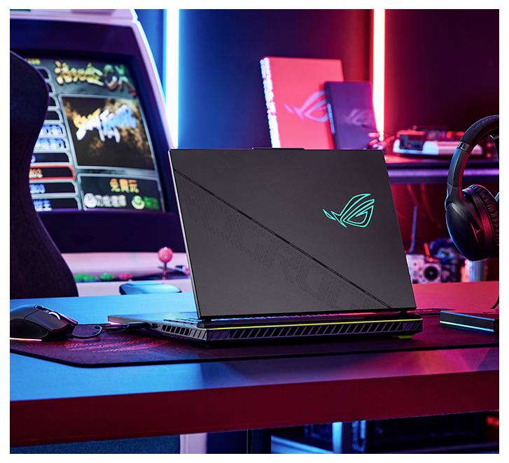 Gaming-Laptop mit beleuchtetem ROG-Logo auf einem Schreibtisch, dargestellt in einem neonbeleuchteten Gaming-Setup mit Arcade-Automat und Headset in der Nähe.