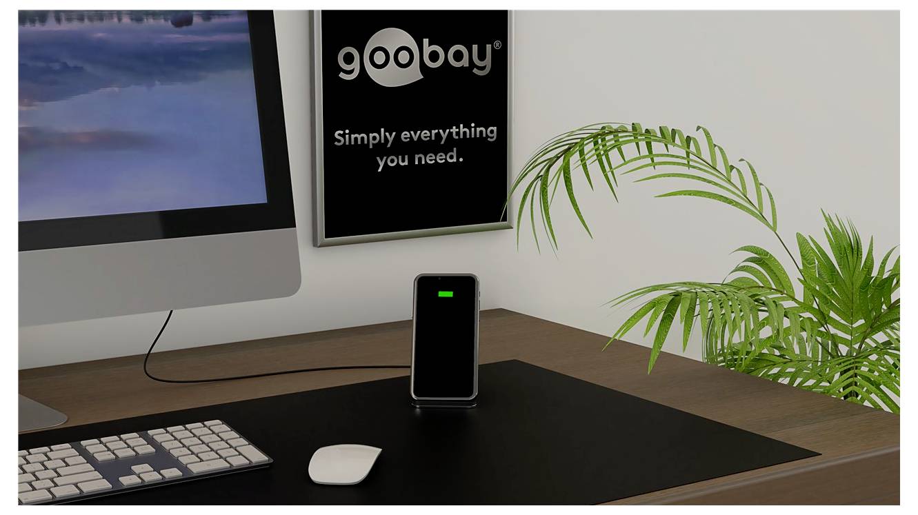 Schreibtischaufbau mit iMac, Tastatur, Maus und einem aufgeladenen Telefon auf einem Ständer neben einem „goobay"-Poster mit dem Text „Einfach alles, was du brauchst."