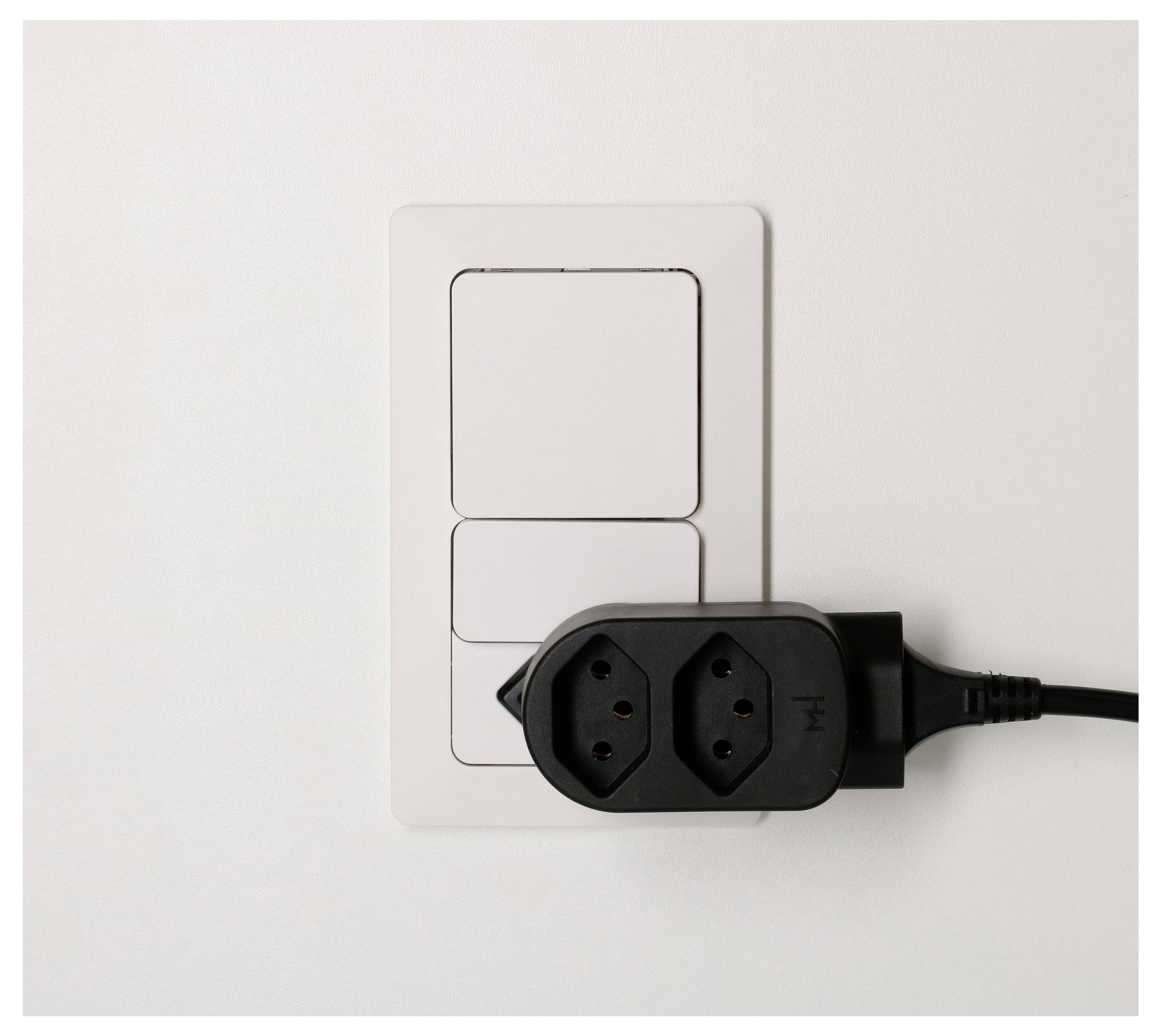 Schwarze Steckdosenleiste mit Stecker, der an eine weiße Wandsteckdosenplatte angeschlossen ist und den unteren Steckdosenbereich abdeckt.