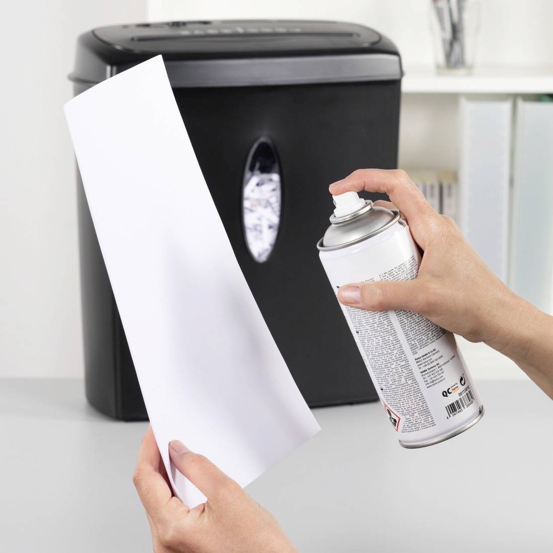 Hände halten ein Blatt Papier und besprühen es mit Aerosol vor einem Aktenvernichter, was auf Reinigung oder Wartung hindeutet.
