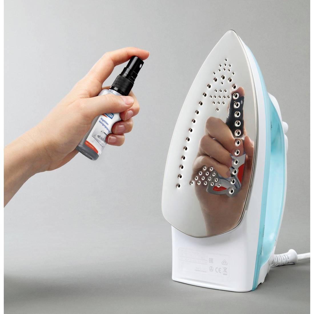 Hand sprays cleaner onto a standing electric iron, suggesting ironing or appliance maintenance.<br><br>---<br><br>Hand sprüht Reiniger auf ein stehendes Elektrobügeleisen, was auf Bügeln oder Wartung von Haushaltsgeräten hindeutet.