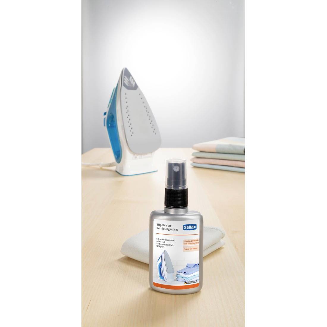 Sprühflasche mit Eisenreiniger auf einem Tisch, mit einem stehenden Dampfbügeleisen und gefalteter Wäsche im Hintergrund.
