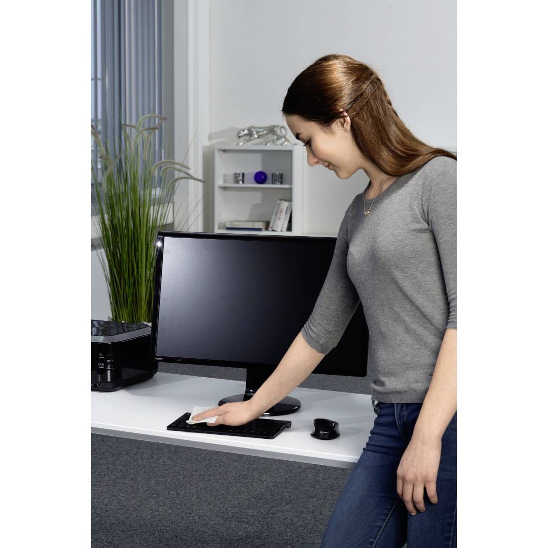 Eine Frau reinigt einen Schreibtisch mit einem Tuch neben einem Computerbildschirm in einem Büro.