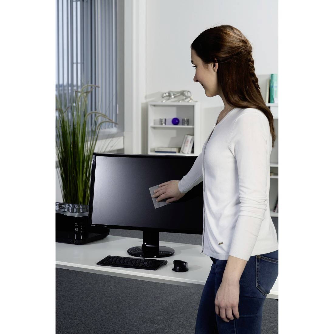Eine Frau reinigt einen Computermonitor mit einem Tuch an einem Schreibtisch im Büro, in der Nähe befinden sich eine Tastatur, eine Maus und eine Pflanze.
