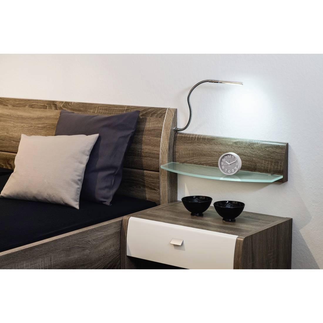 Moderne Schlafzimmerecke mit einem Holzbett, Kissen, wandmontiertem Regal und Lampe, kleine Uhr und Nachttisch mit Schüsseln.