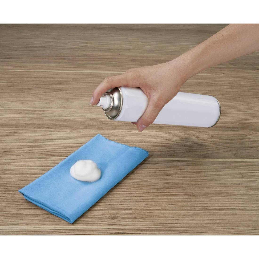 Mit der Hand wird weißer Schaumreiniger auf ein blaues Tuch auf einem Holztisch gesprüht.