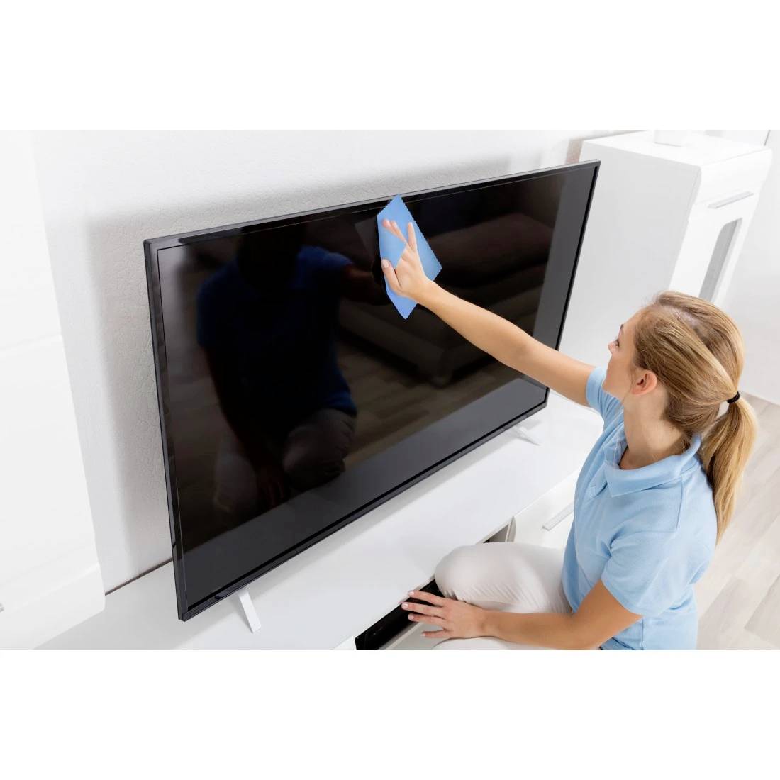 Frau, die einen Flachbildfernseher mit einem blauen Tuch in einem hellen Wohnzimmer reinigt