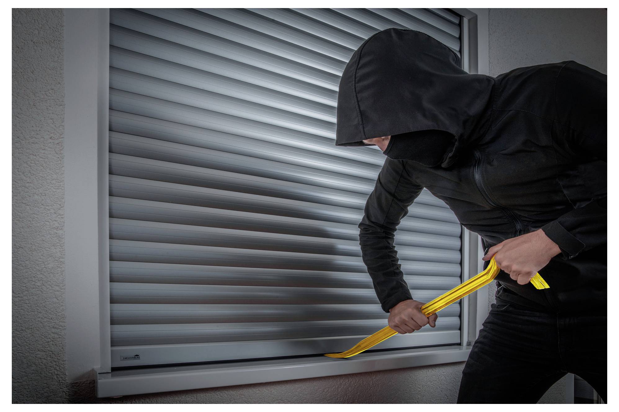 Ein maskierter Einbrecher hebelt mit einer gelben Brechstange ein Fensterrolladen auf, was auf einen Einbruchsversuch hindeutet.
