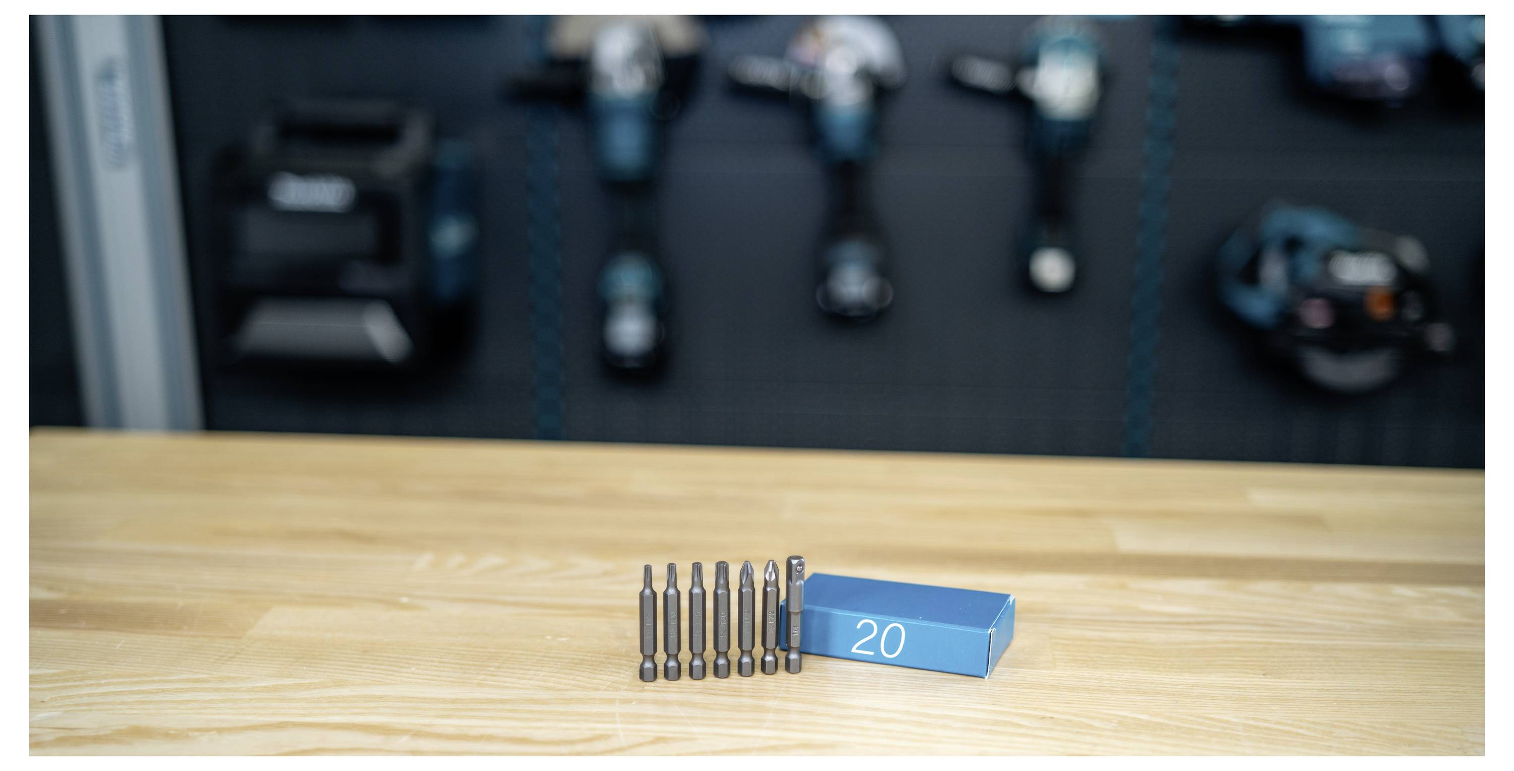 Sechs Bohrer in einer Reihe neben einer blauen Schachtung mit der Aufschrift „20" auf einem Holztisch, mit unscharfem Werkstatt-Hintergrund.