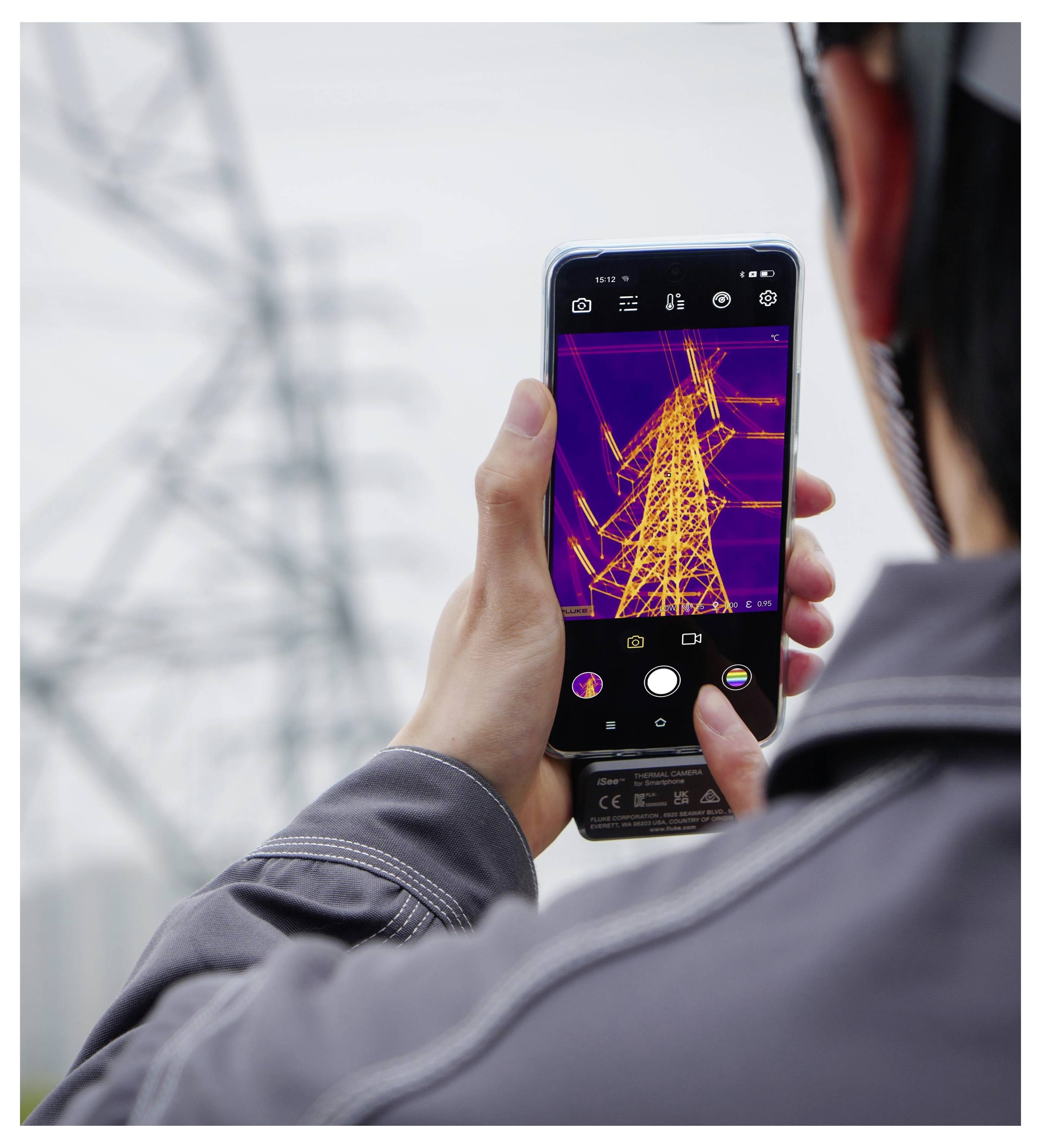 Eine Person fotografiert einen Stromübertragungsmast mit einem Smartphone; der Bildschirm zeigt den Mast in leuchtenden thermischen Farben gegen einen unscharfen Außenhintergrund.
