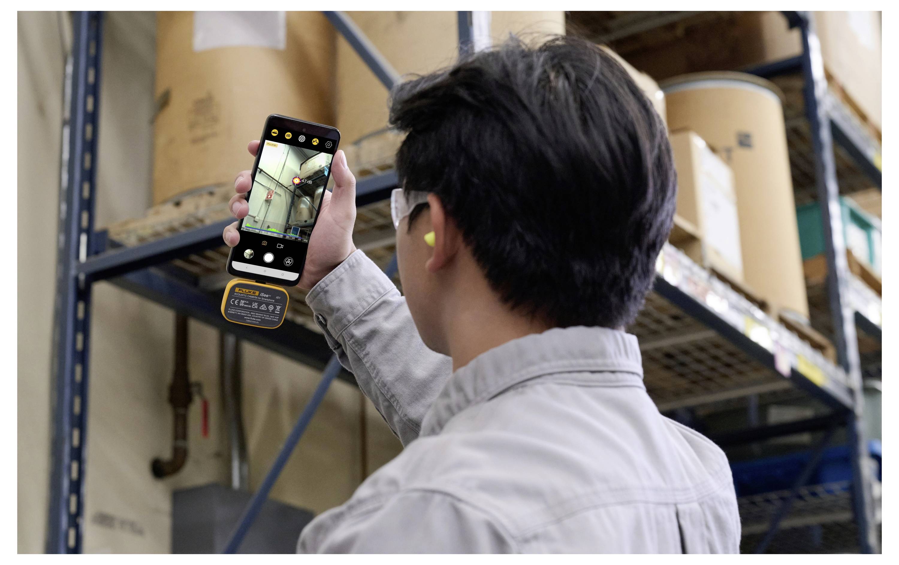 Arbeiter, der ein Smartphone nutzt, um Lagorregalfächer und gestapelte Kartonschachteln zu inspizieren.