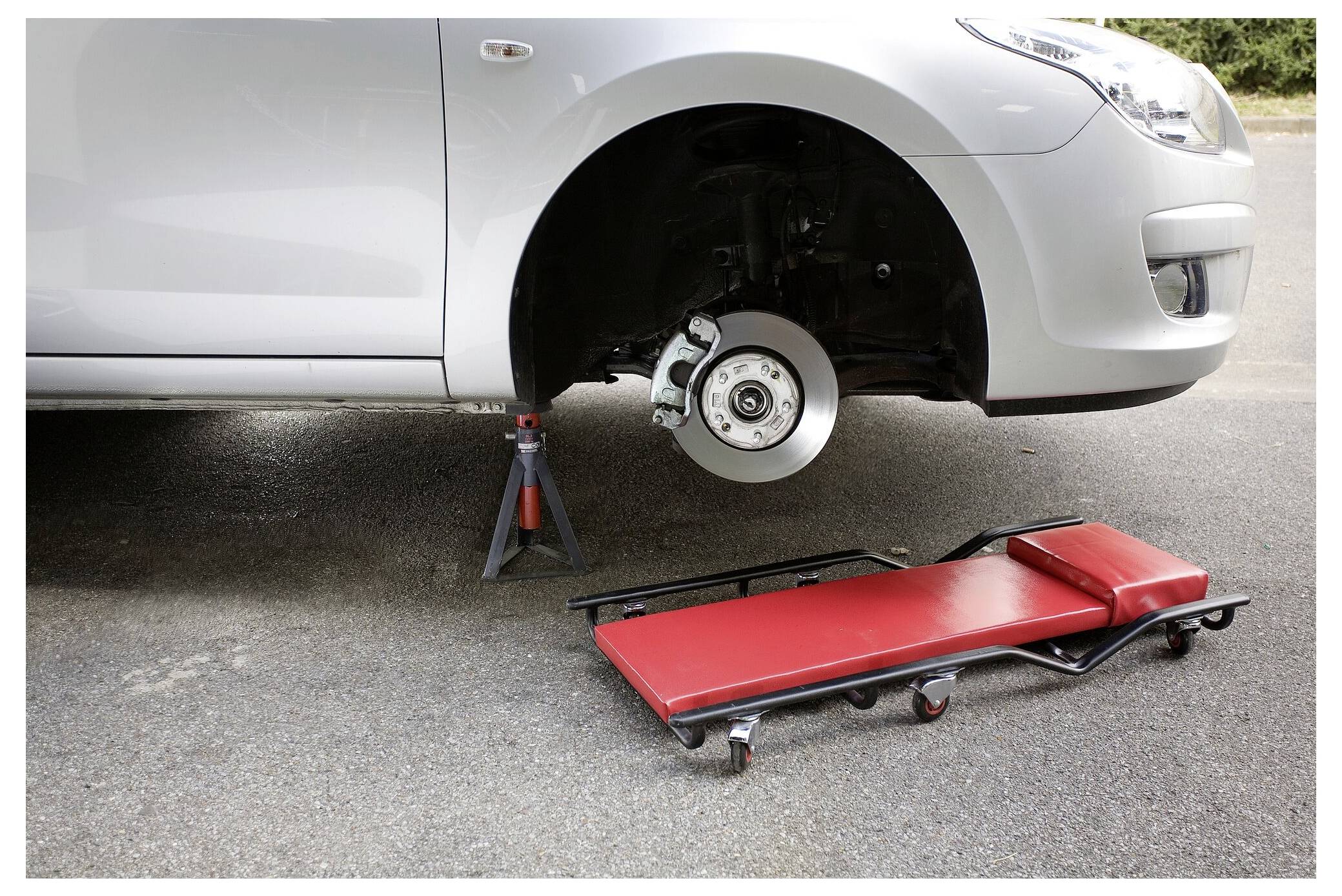 Auto auf Böcken mit entferntem Vorderrad und einer roten fahrbaren Mechaniker-Liege darunter, was auf Fahzeugreparatur oder Wartung hindeutet.