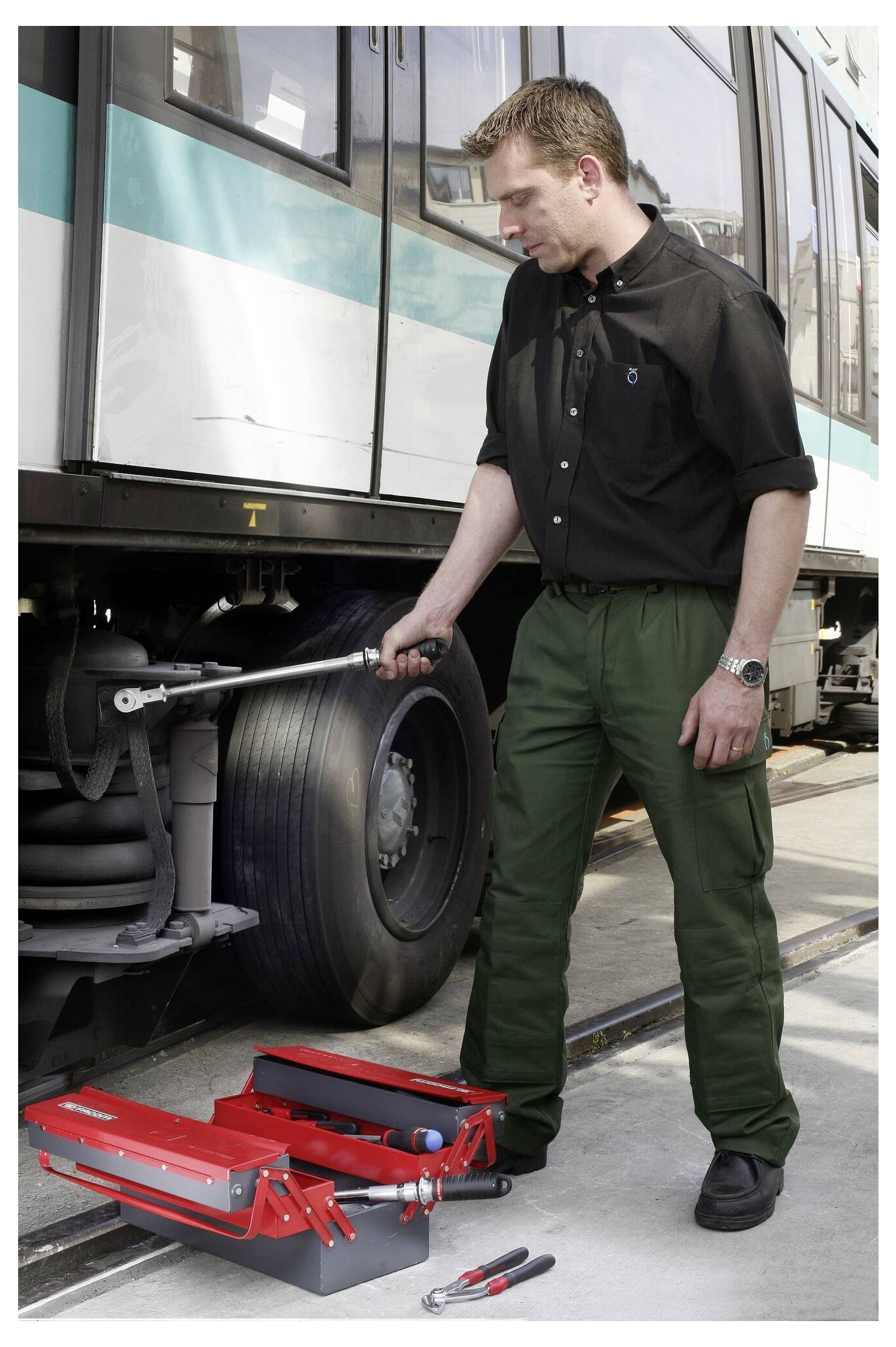 Ein Mann inspiziert ein Busrad mit einem roten Spannwerkzeug auf dem Boden neben einer Zange, wahrscheinlich zur Vorbereitung von Reifen- oder Radwartung.