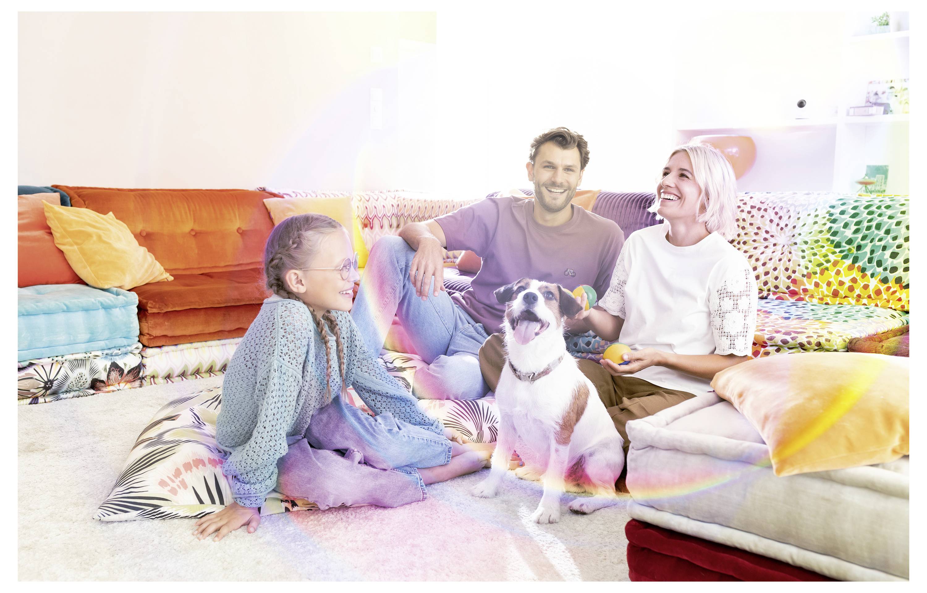 Familie sitzt auf dem Wohnzimmerboden mit einem Hund, lächelt zusammen auf bunten Kissen und einem Sofa.