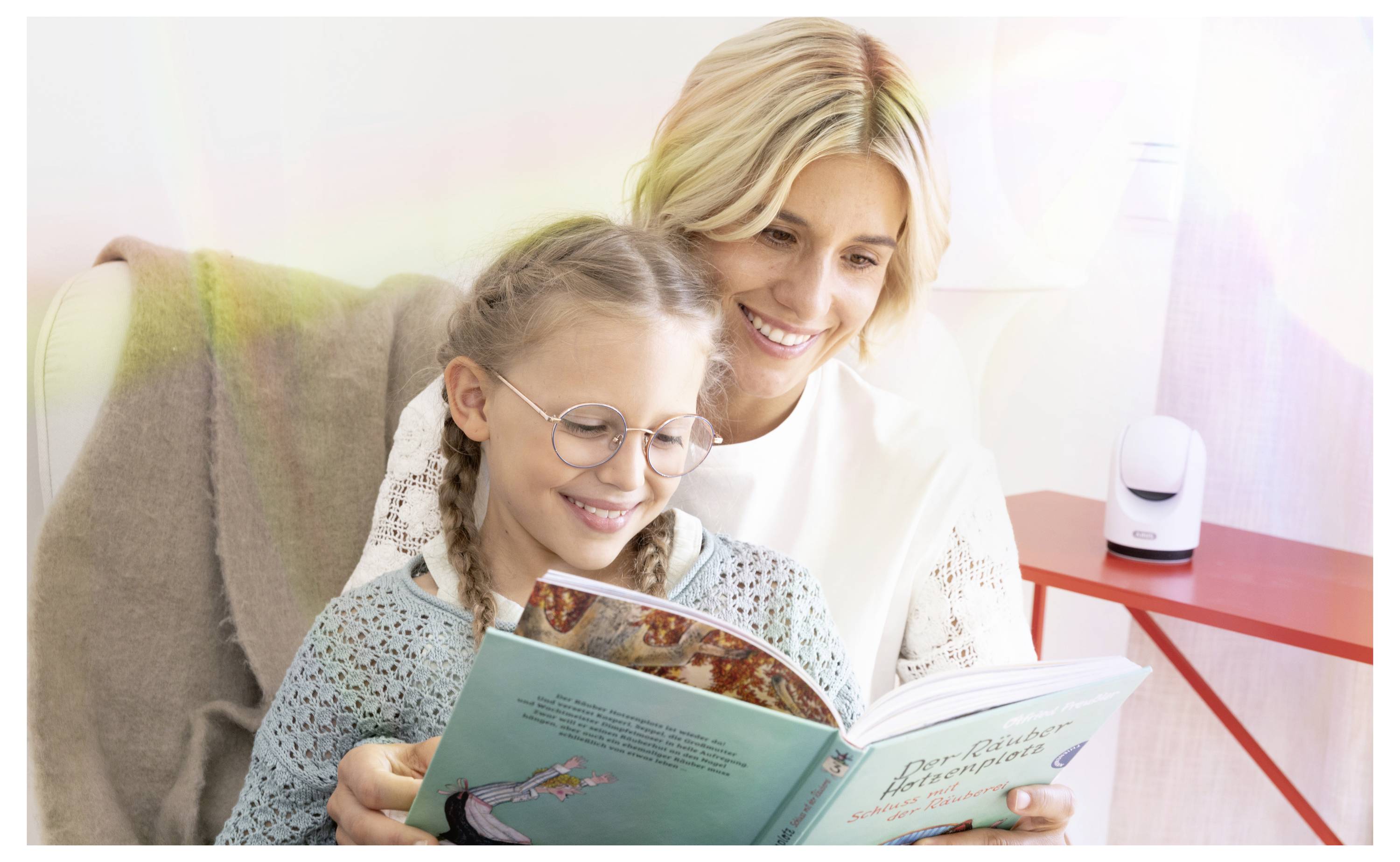 Eine lächelnde Frau und ein junges Mädchen lesen zusammen ein Buch auf einem Sofa in einem hellen Wohnzimmer.