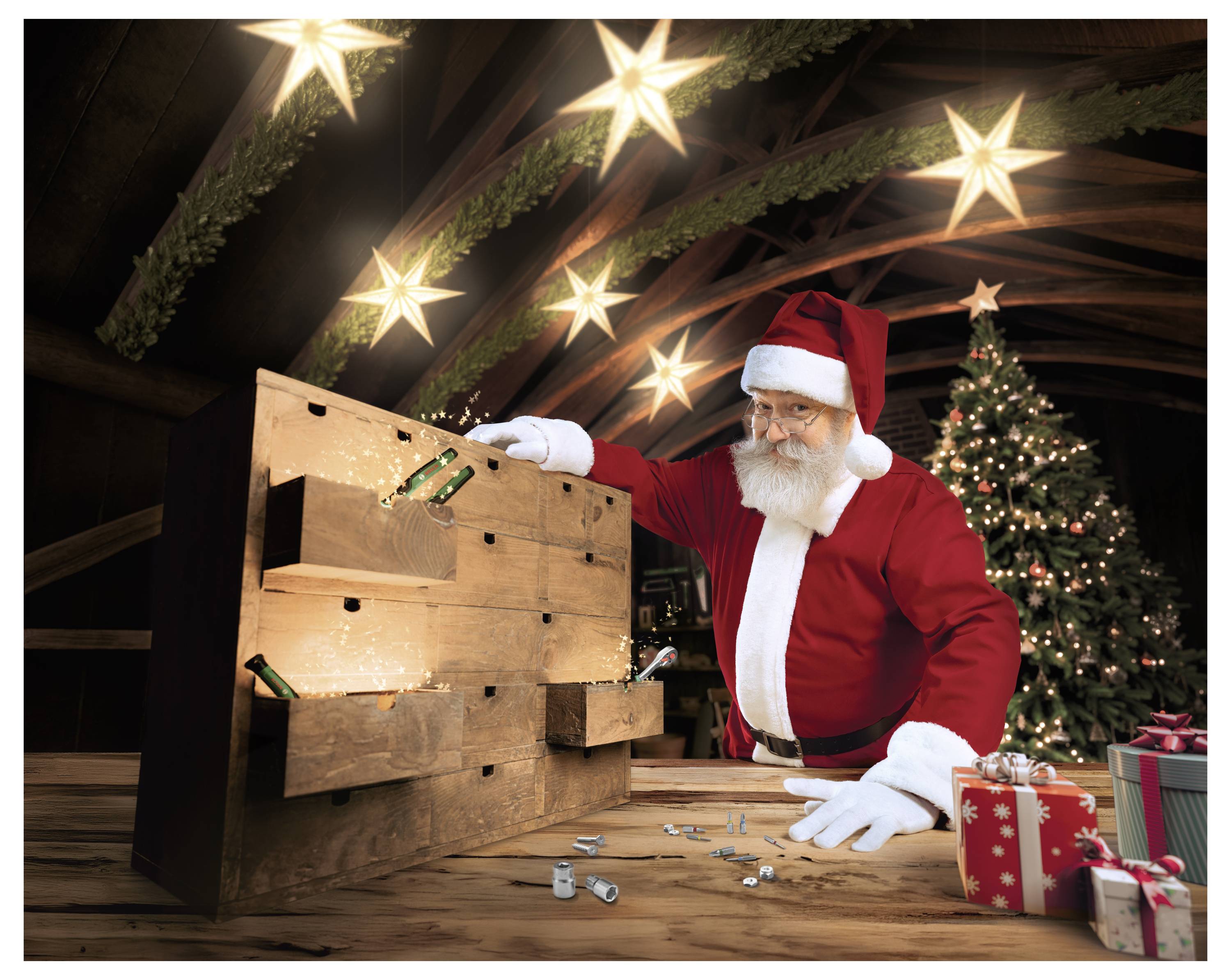 Der Weihnachtsmann beugt sich über eine Holztruhe mit Schubladen in einer Werkstatt, mit Weihnachtssternen über ihm und einem geschmückten Baum sowie Geschenken im Hintergrund.